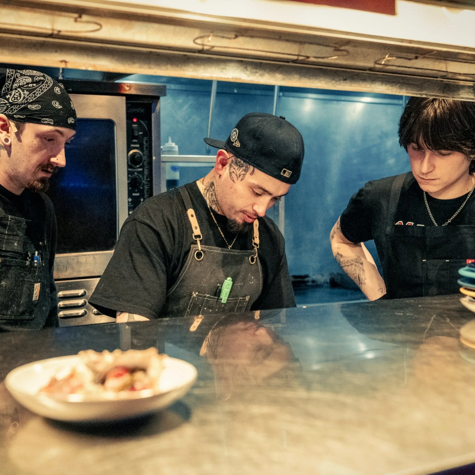 Three male chefs working in a restaurant kitchen, viewed from below the counter, with a bowl of pizza in the foreground.