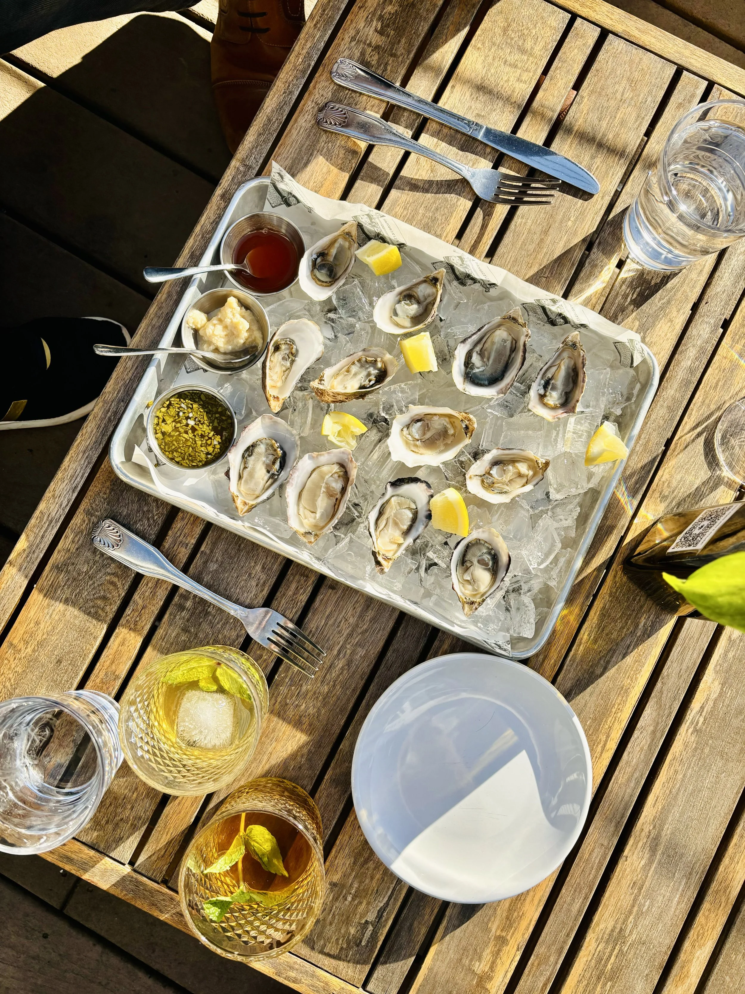 A wooden outdoor table with a tray of cooked oysters on ice, small bowls of sauces and lemon wedges, forks and knives, and glasses of water, with sunlight and shadows.