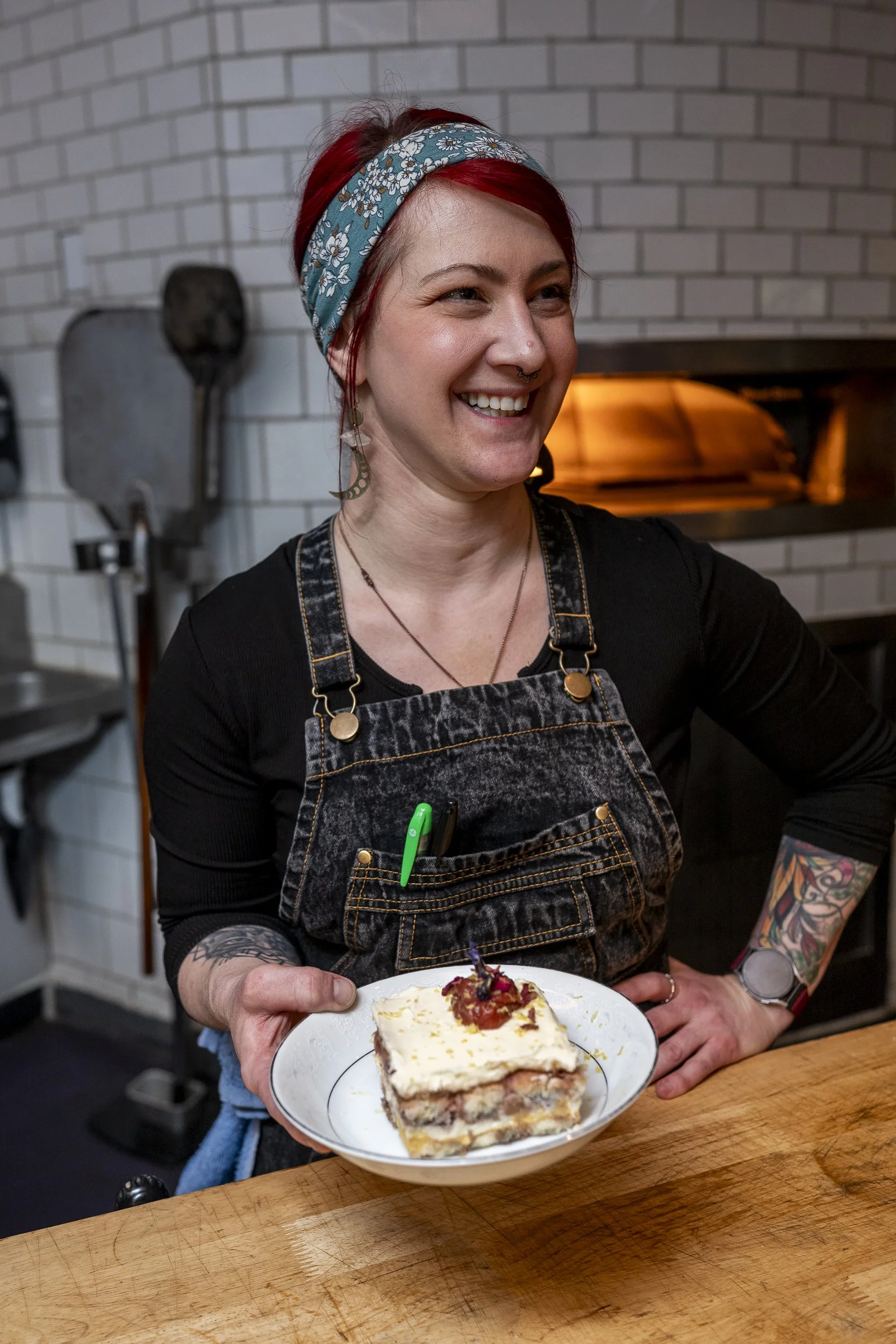 A woman with red hair, a floral headband, and tattoos on her arms, holding a plate with a slice of layered cake topped with fruit, standing in a kitchen with brick walls and oven.
