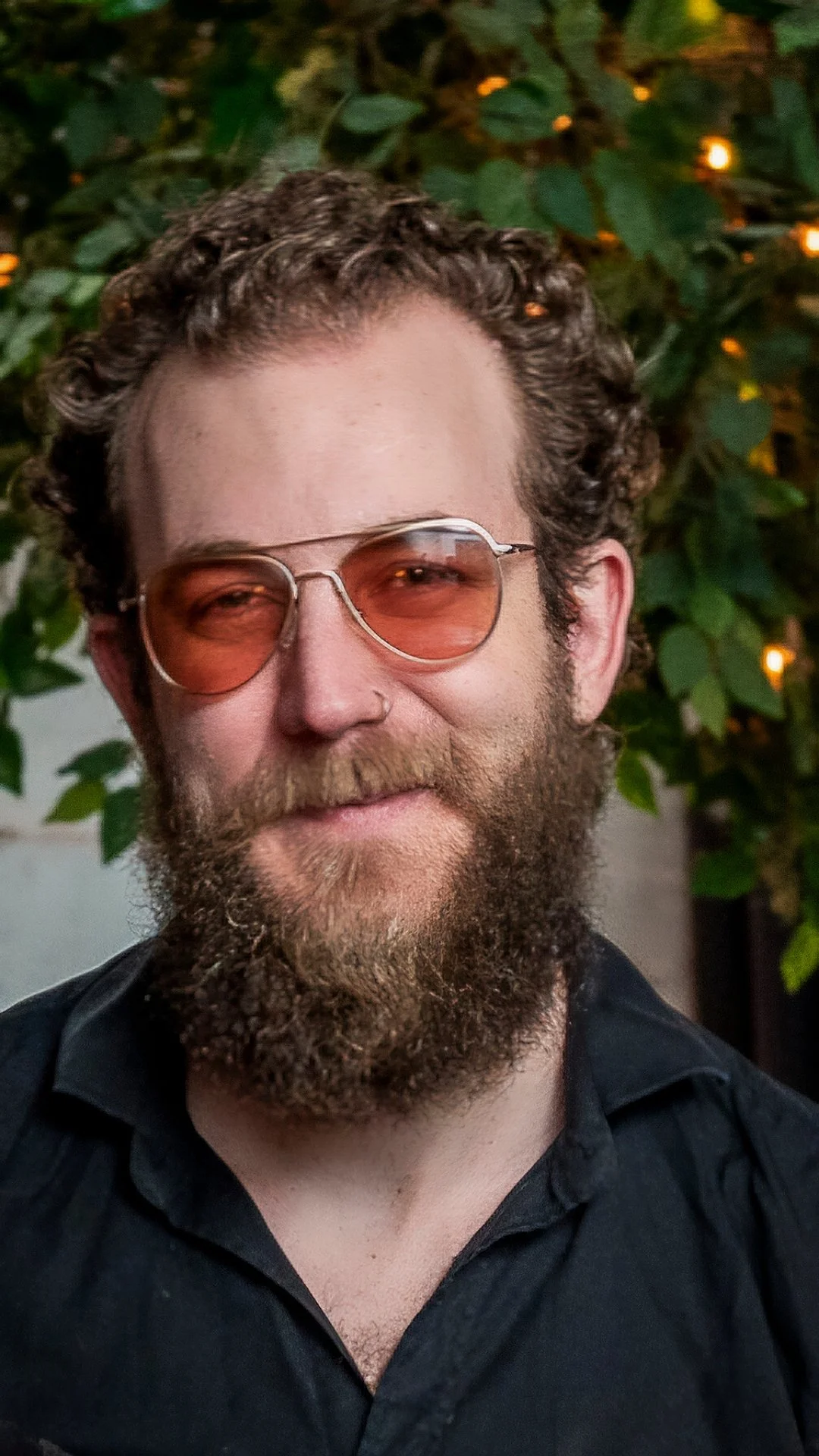A man with curly hair, a beard, and glasses with orange-tinted lenses smiling in front of green leafy plants with some small lights.