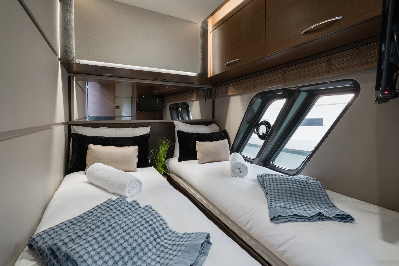 Interior of a bedroom with two beds, each with rolled towels, pillows, and blankets, located inside a boat or yacht with porthole windows and wooden cabinets.