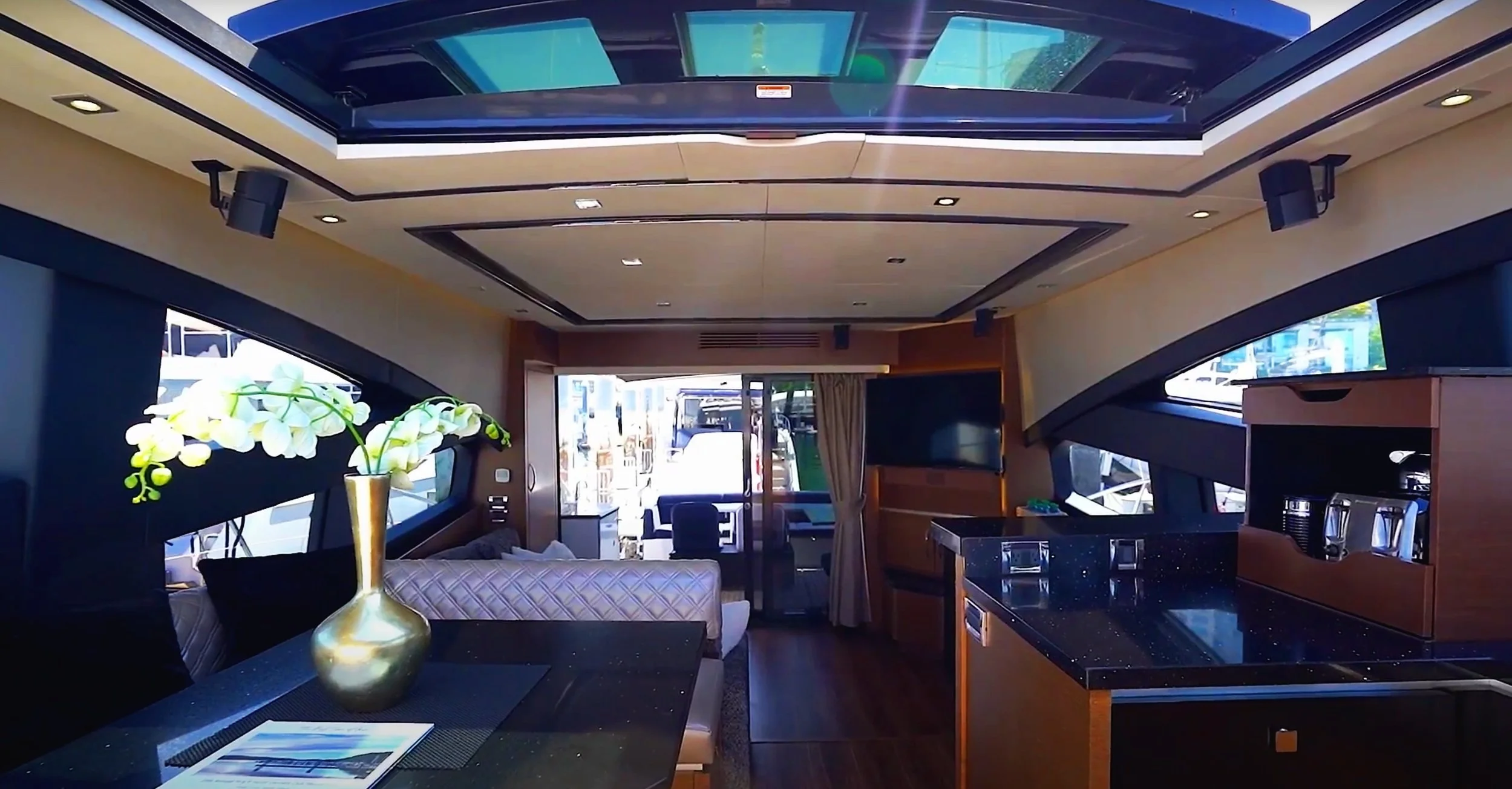 Interior of a boat or yacht with a modern design, featuring a ceiling with skylights, large windows, a dining area with a vase of white flowers, and a small kitchen or bar area.