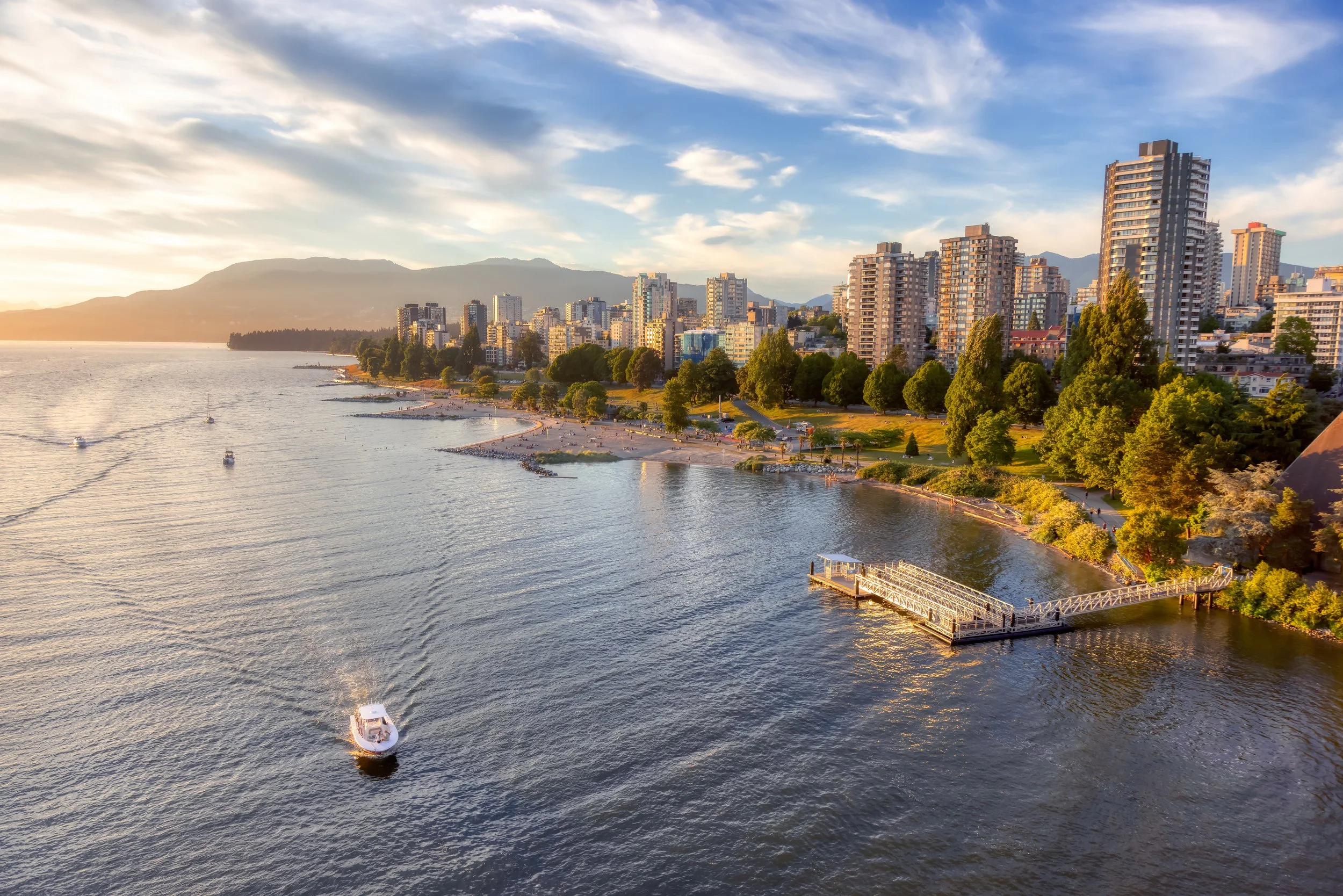 City skyline with tall buildings along a waterfront during sunset, with boats on the water and a pier extending into the river.