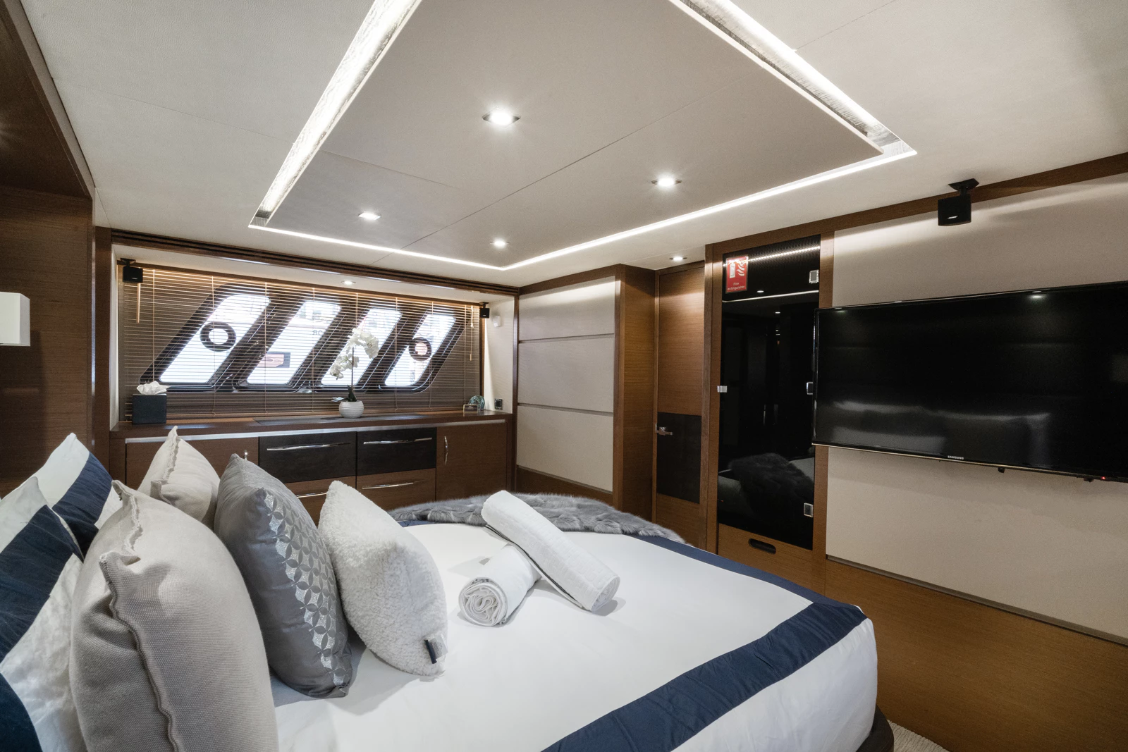 Modern bedroom with a bed covered in white linens and decorative pillows, rolled towels on the bed, a large flat-screen TV on the wall, a window with blinds and angled skylights, and warm wood paneling.