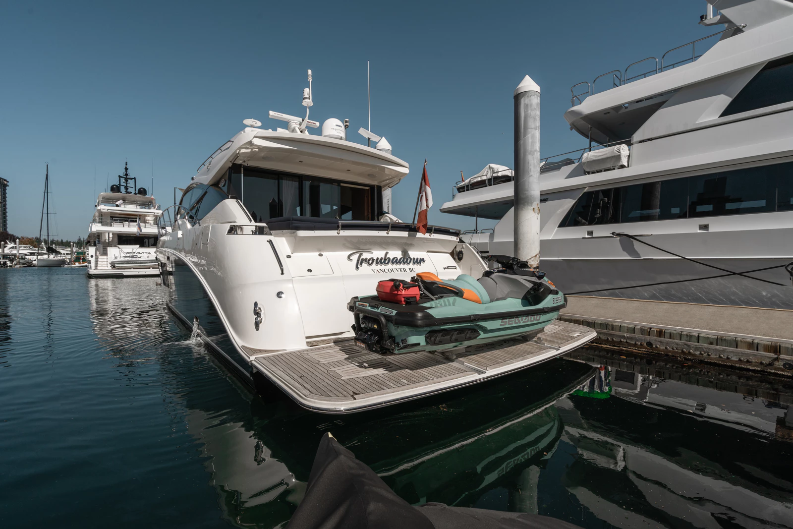 A marina scene with several white yachts docked, including one with the name 'Troubadour' in Vancouver BC. A small green Sea-Doo jet ski is on the rear swim platform of the Troubadour, with a red fuel container on top. The water is calm and the sky i