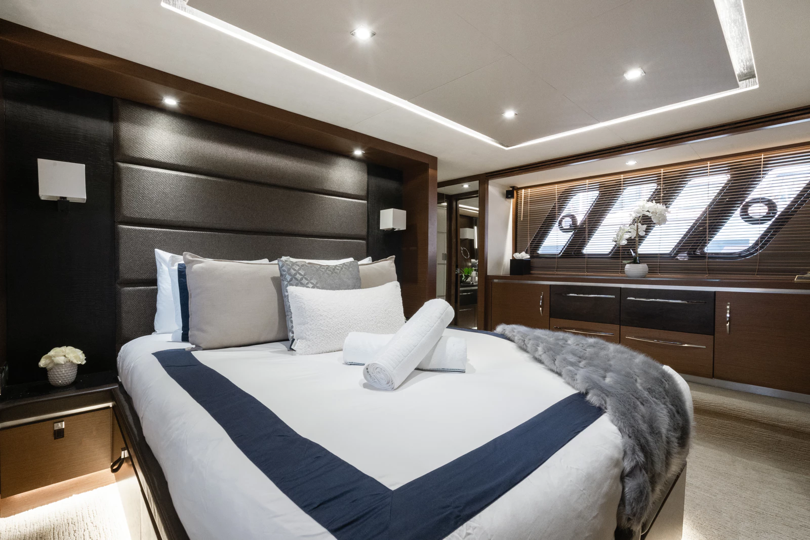 Luxurious bedroom with a large bed, decorative pillows, and rolled towels. The room features modern lighting, a padded headboard, wooden accents, and a row of windows with venetian blinds and a flower vase.
