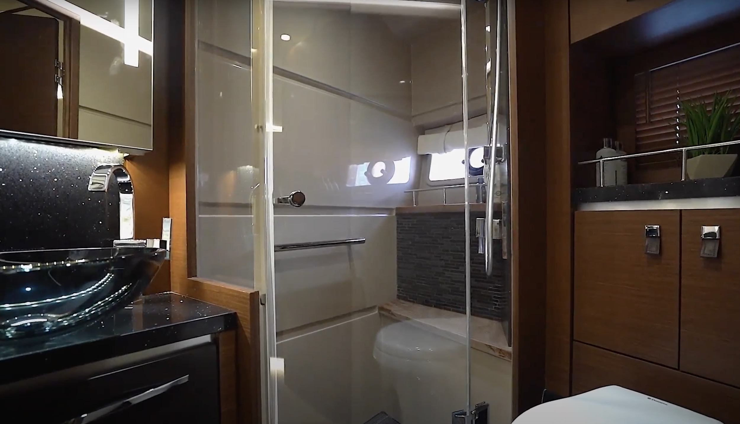 Interior of a bathroom with a shower enclosure, wooden cabinets, a window with blinds, a sink, and a black countertop with a stovetop in the corner.