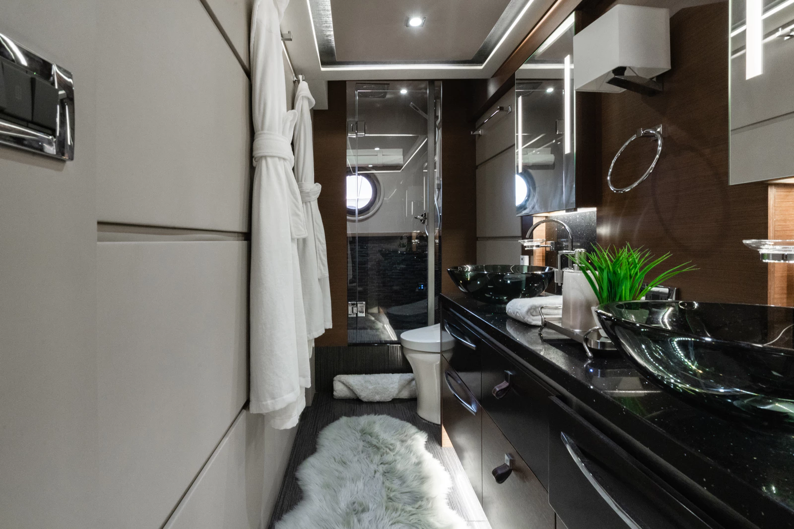 Luxurious bathroom featuring black vessel sinks on a black countertop, a glass-walled shower, and beige cabinetry with a window in the shower area. Decor includes a white bathrobe, fluffy white rug, green plant, and modern fixtures.