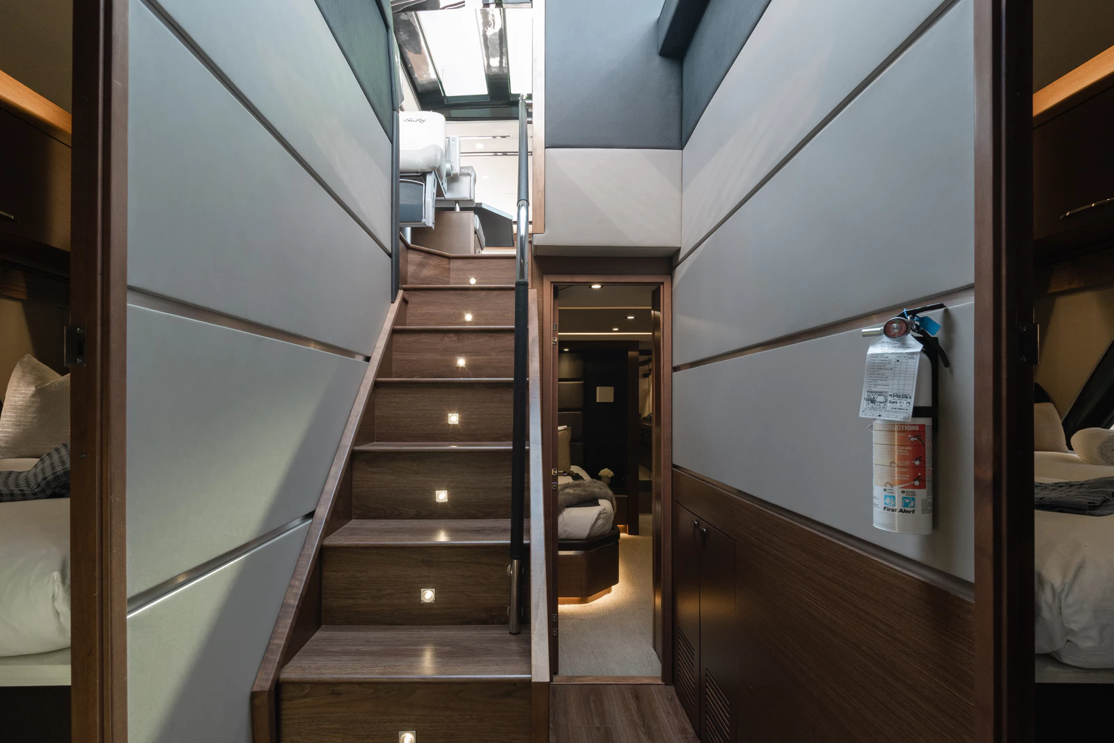 Inside a modern small living space with a staircase with built-in lights leading to an upper level. Visible areas include a doorway, a fire extinguisher on the wall, and part of a bedroom with a bed and pillows.