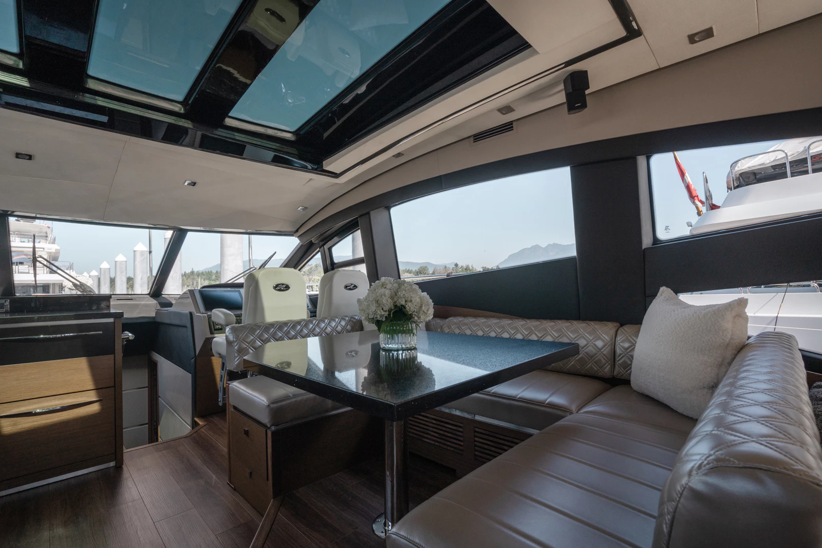 Interior of a yacht with a dining area, including a black table with a vase of white flowers, beige cushioned seating, and a panoramic window view of the water and other yachts.