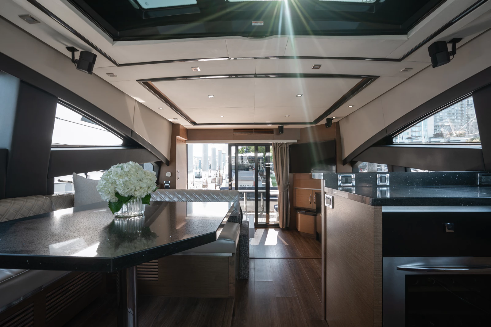 Interior of a modern yacht with a dining area, a glass vase with white flowers on the table, and a view of a marina through sliding glass doors.