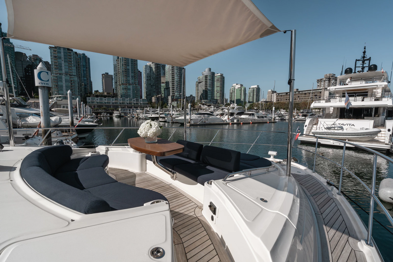 Luxury yacht deck with outdoor seating, a wooden table with a flower bouquet, and cityscape with high-rise buildings and boats in the harbor in the background.
