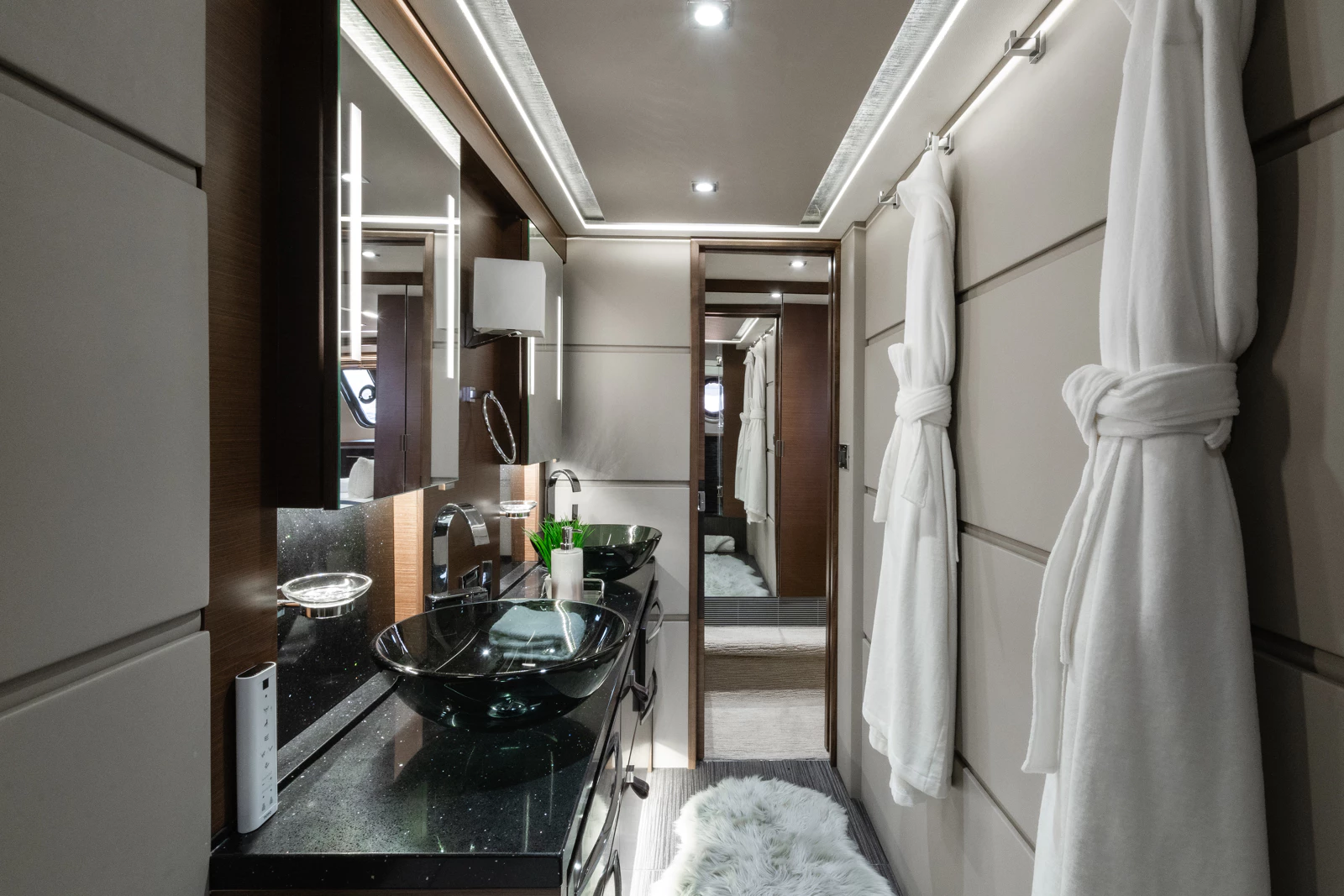 A modern bathroom with two black glass vessel sinks on a black countertop, round mirrors, white bathrobes hanging on a wall, a fluffy white rug on the floor, and a doorway leading to a bedroom.