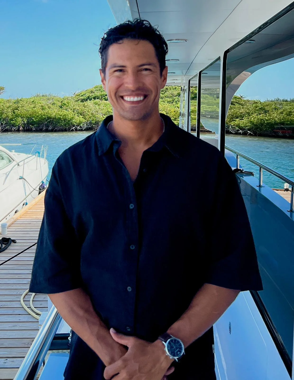 Smiling man in black shirt standing on a boat dock with water and green trees in the background.