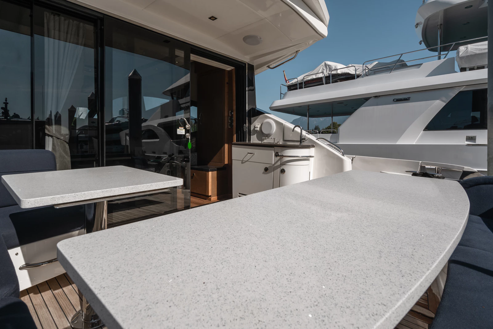 Outdoor dining area on a yacht with two light-colored tables and dark cushioned chairs, with yacht and docked boats in the background under a clear sky.