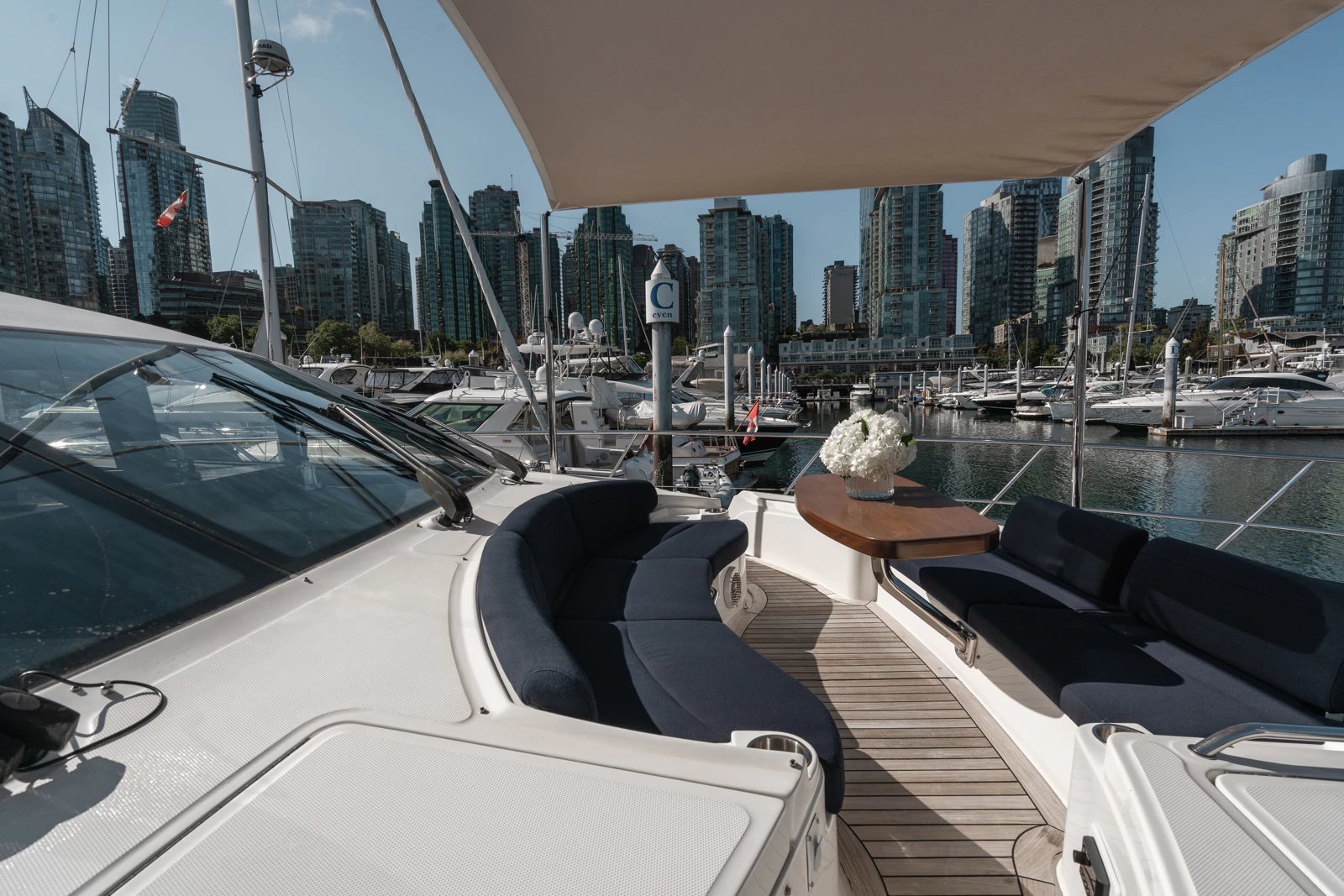 View of a yacht deck with navy cushioned seating, a wooden table with a flower arrangement, overlooking a marina with yachts and skyscrapers in the background.