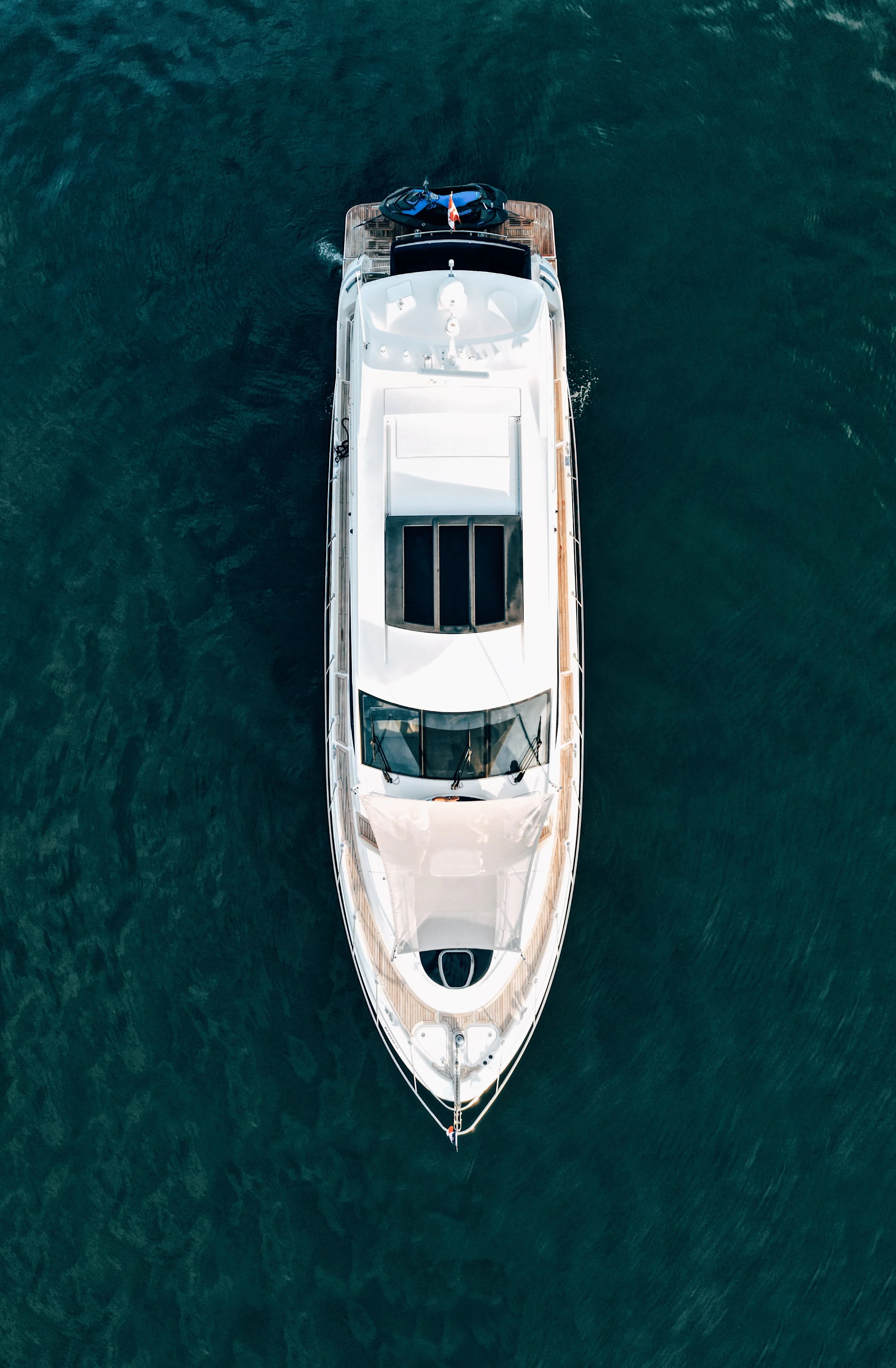 Overhead view of a white yacht sailing in dark green waters, with a smaller boat attached at the stern and a Canadian flag flying at the stern.