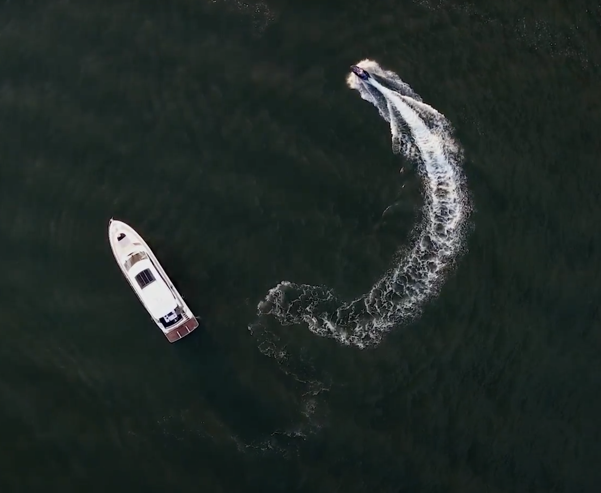 Overhead view of a motorboat and a jet ski on dark green water, with the boat creating a wake.