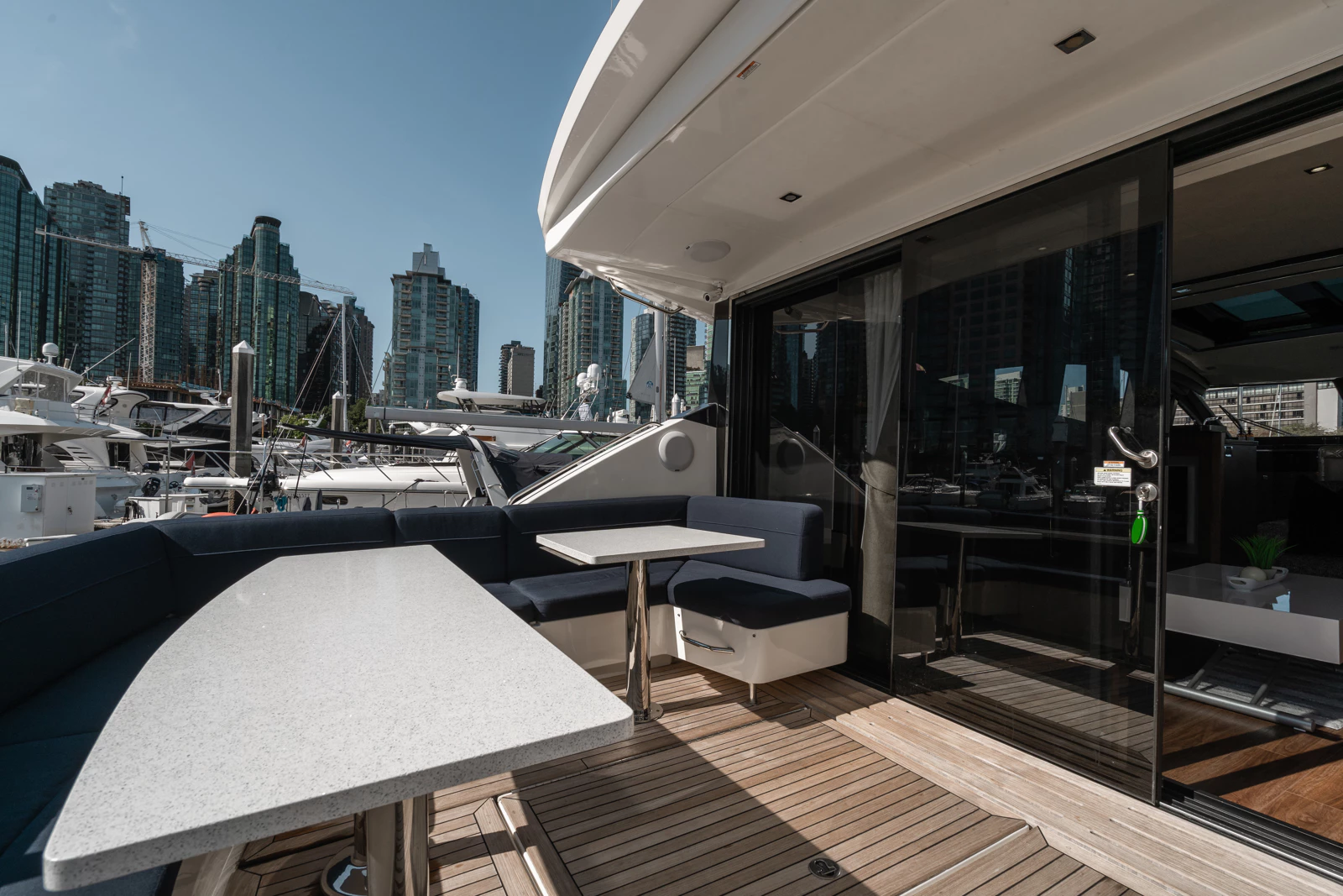 Outdoor deck area of a yacht with a table, cushioned seating, and sliding glass door leading inside, with marina and city skyline in the background.