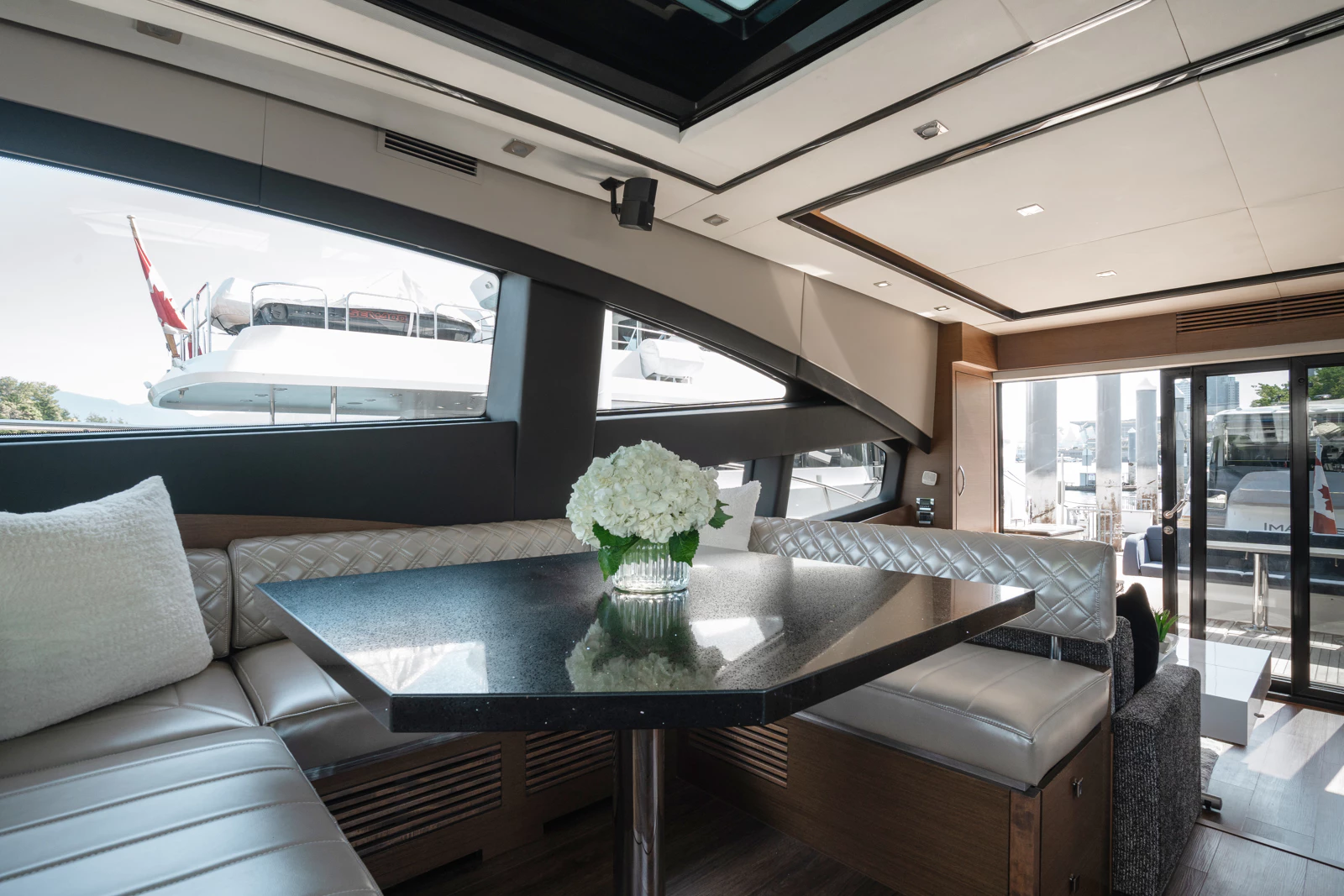 Interior of a yacht with a black countertop table, white cushioned seating, a vase with white flowers, and large windows showing docked boats outside.