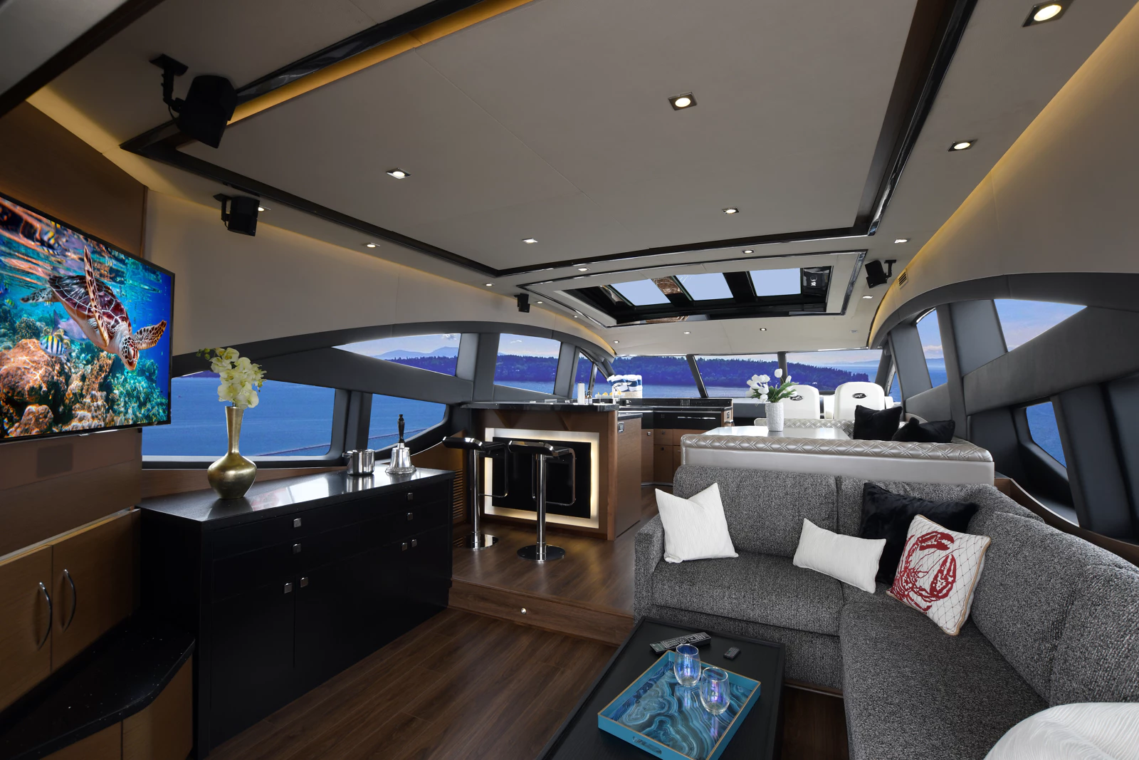 Interior of a modern yacht with large windows showing water and mountains, a flat-screen TV displaying a sea turtle, a gray sectional sofa with decorative pillows, a black cabinet with a vase and flowers, and a bar area with stools.