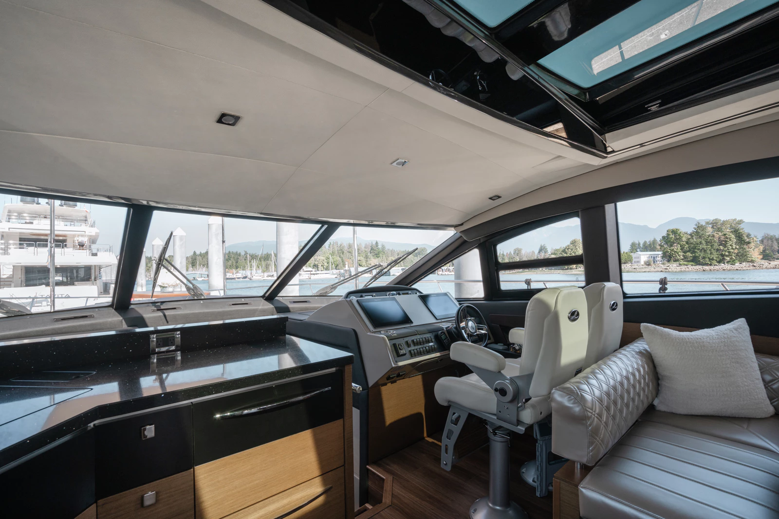 Interior of a yacht's bridge with navigation screens, steering wheel, and beige seating, overlooking a marina with boats and water, mountains, and sky in the background.