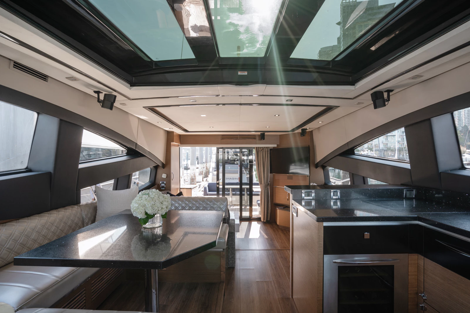 Interior of a yacht with a dining table, white flowers, leather seating, large windows, a television, and a kitchen area.