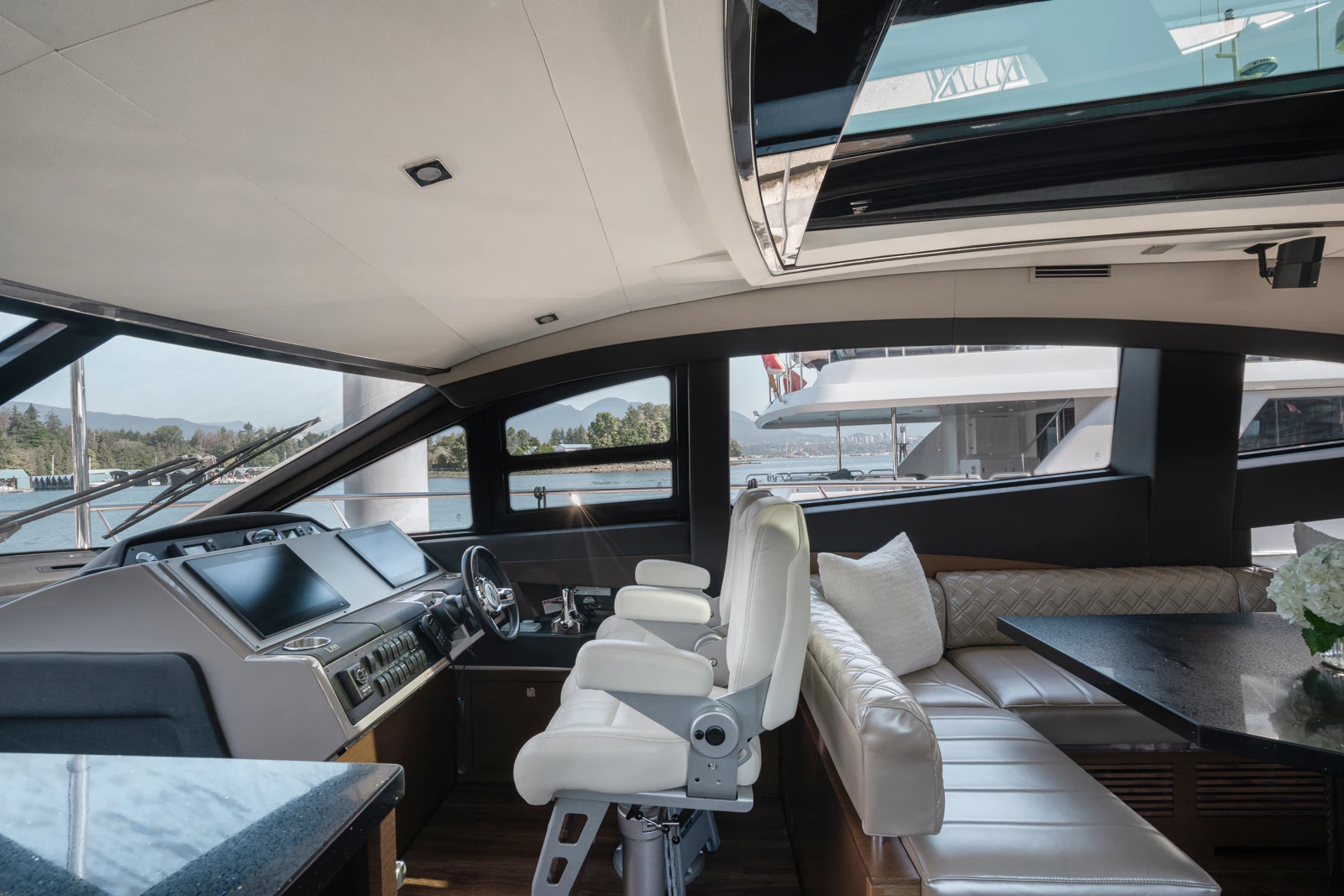 Inside of a yacht featuring a control panel with screens, a white captain's chair, a beige leather sofa with pillows, a black dining table, and large windows showing water and other boats outside.