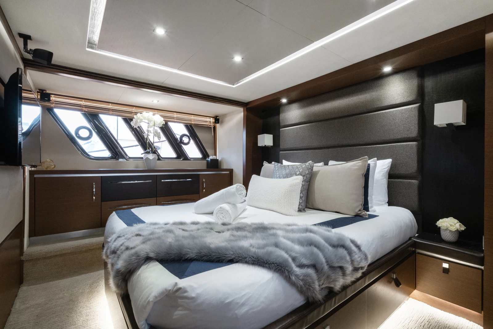 Modern bedroom with a bed dressed in white linens, grey and beige pillows, a grey faux fur throw, placed against a black and grey padded headboard, with a window featuring angled black frames and wooden blinds, decorated with white flowers in vases o