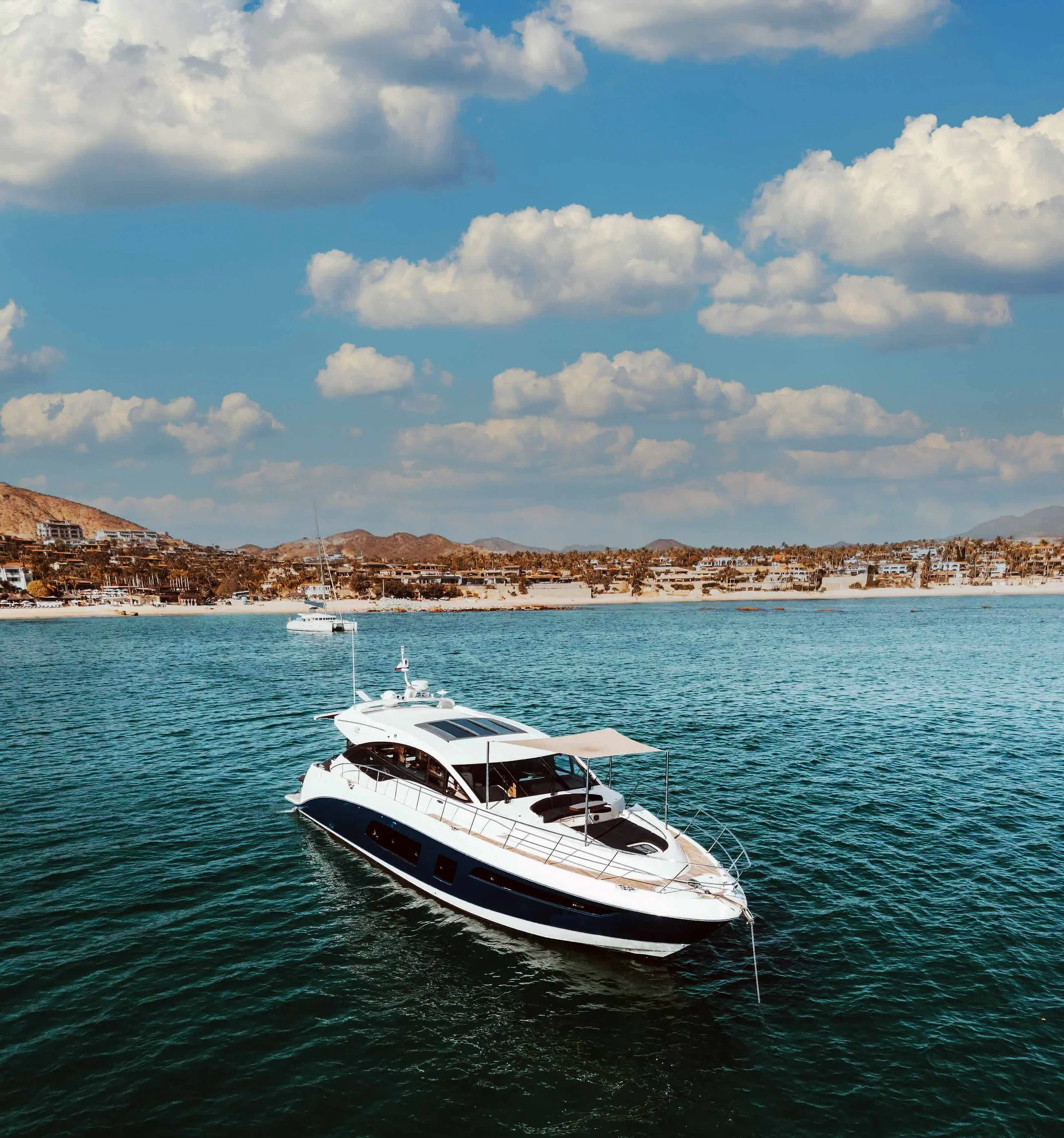 A white yacht floating on turquoise water with a coastline of residential buildings and hills under a partly cloudy sky.