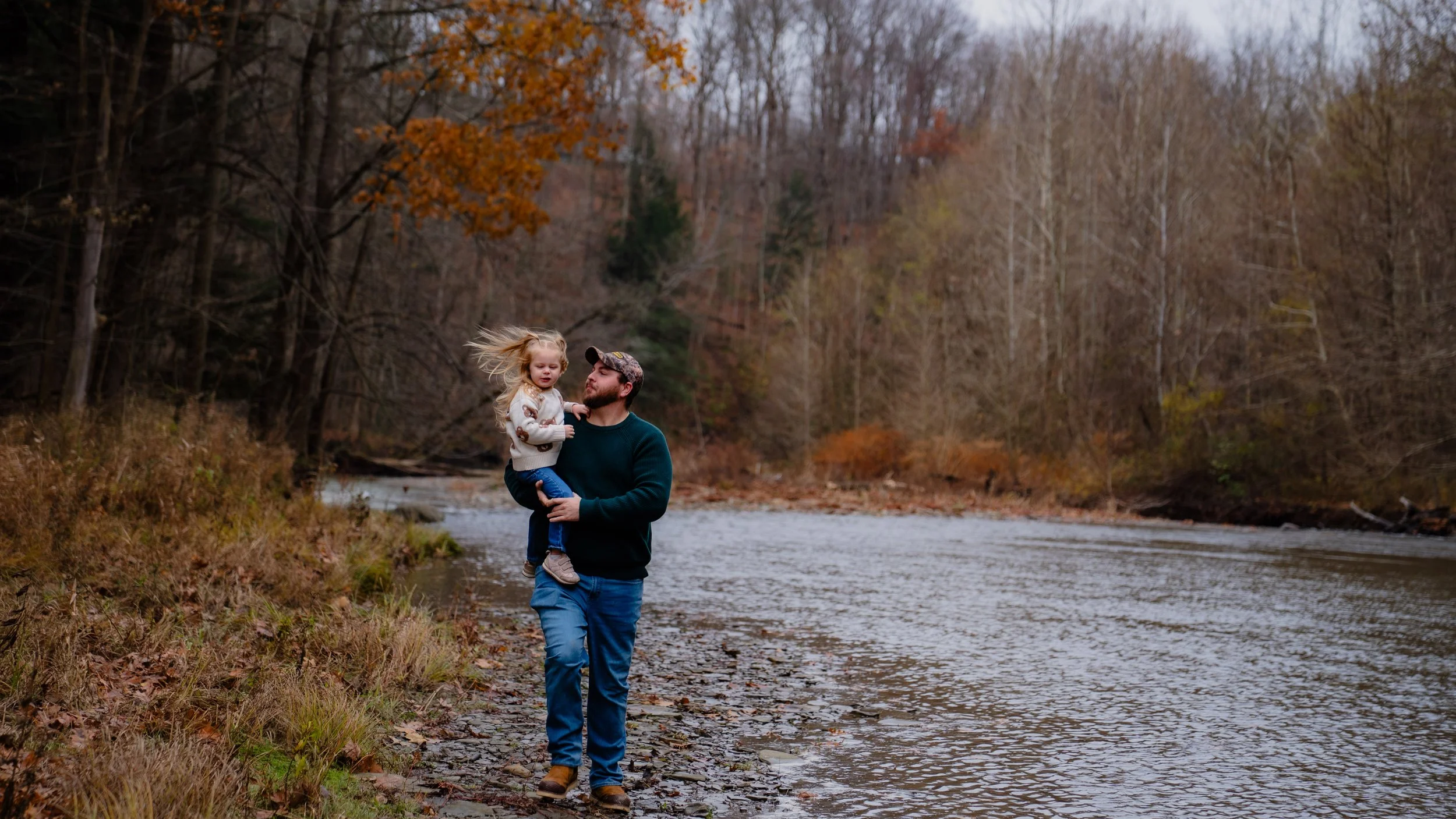 A man holding a young girl in his arms while walking along a riverbank surrounded by trees with autumn foliage.