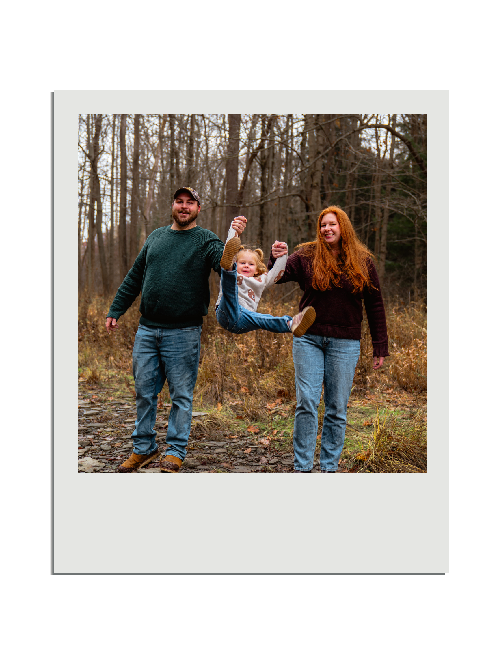 A family of three, a man and woman, are holding a little girl by her hands as she hangs in the air, smiling in a forest during autumn.