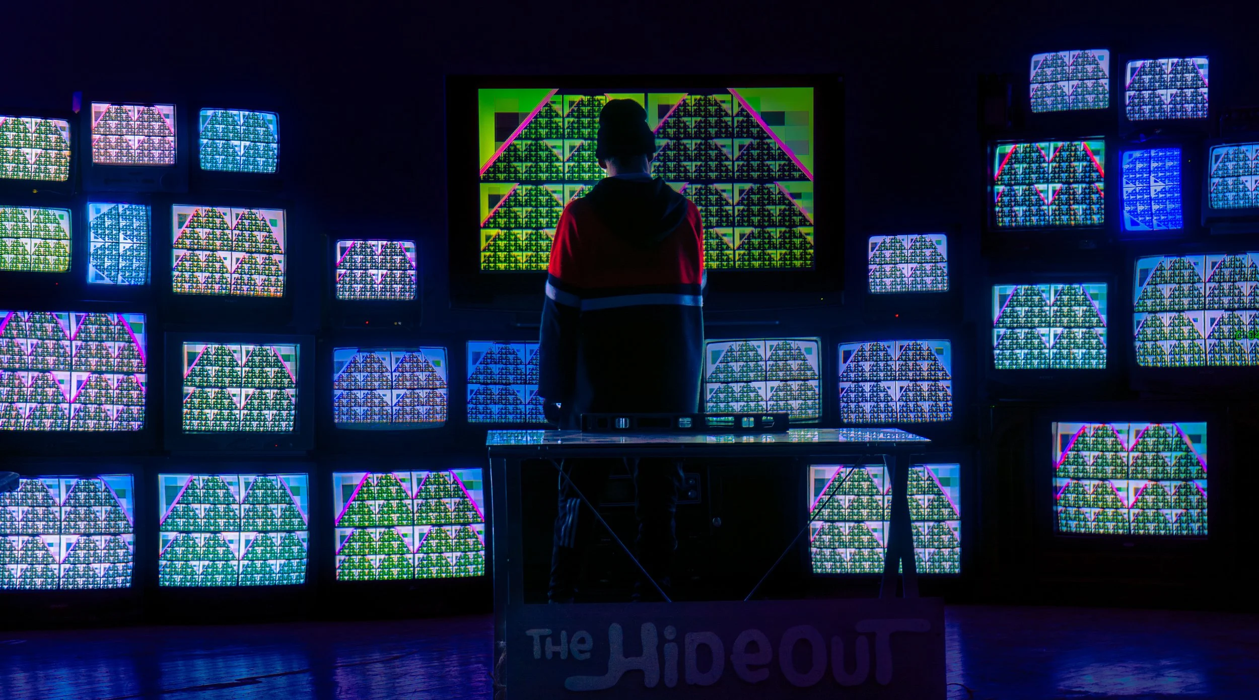 Person wearing a red and black jacket standing in front of a large screen with colorful abstract digital patterns, surrounded by multiple smaller screens displaying similar digital visuals, in a dark room with a sign reading 'The Hideout' on a table.
