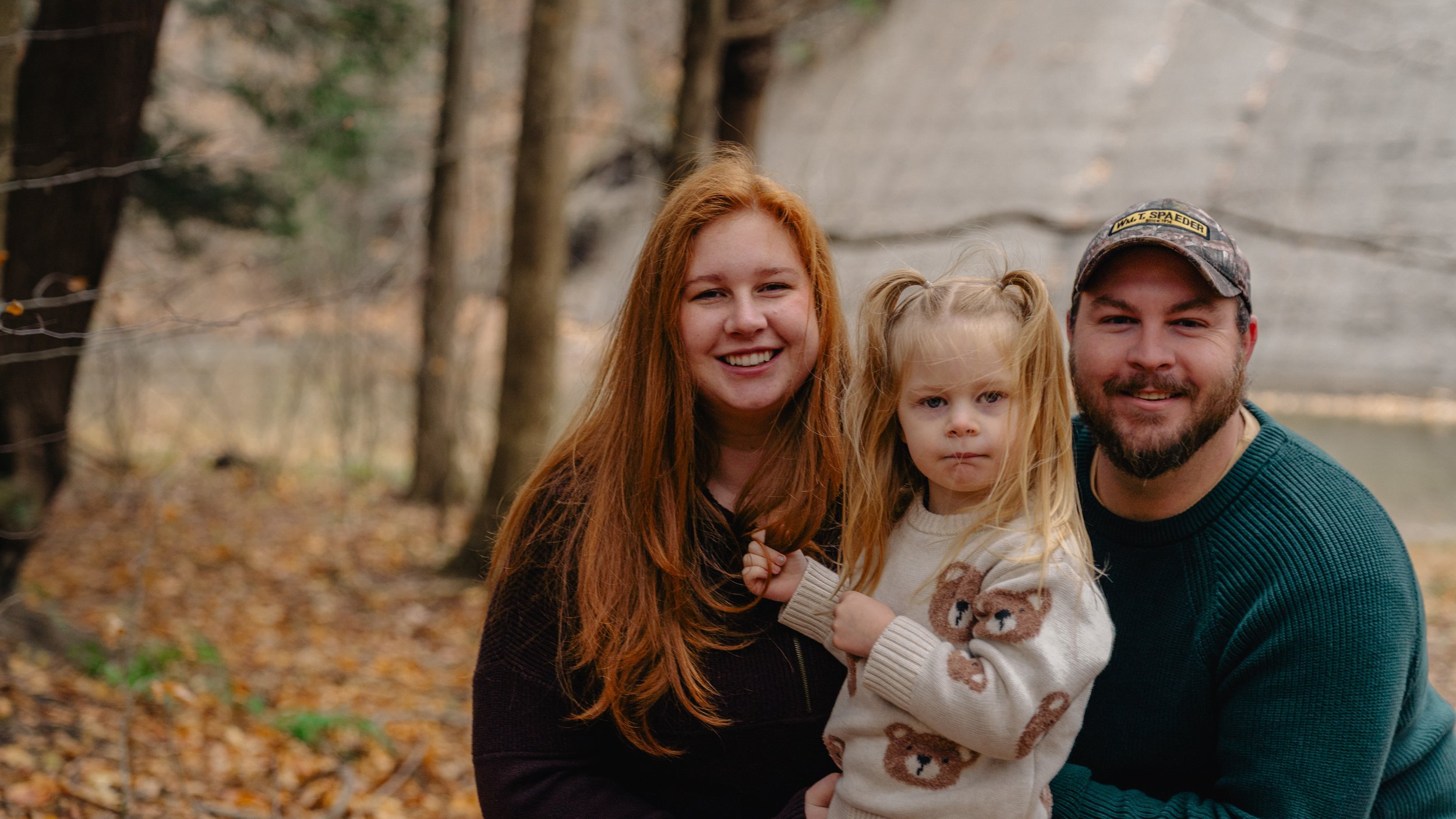 A smiling woman with long red hair, a young girl with blonde hair and a man with a beard, all outdoors in a wooded area during fall, posing with the girl in the middle.