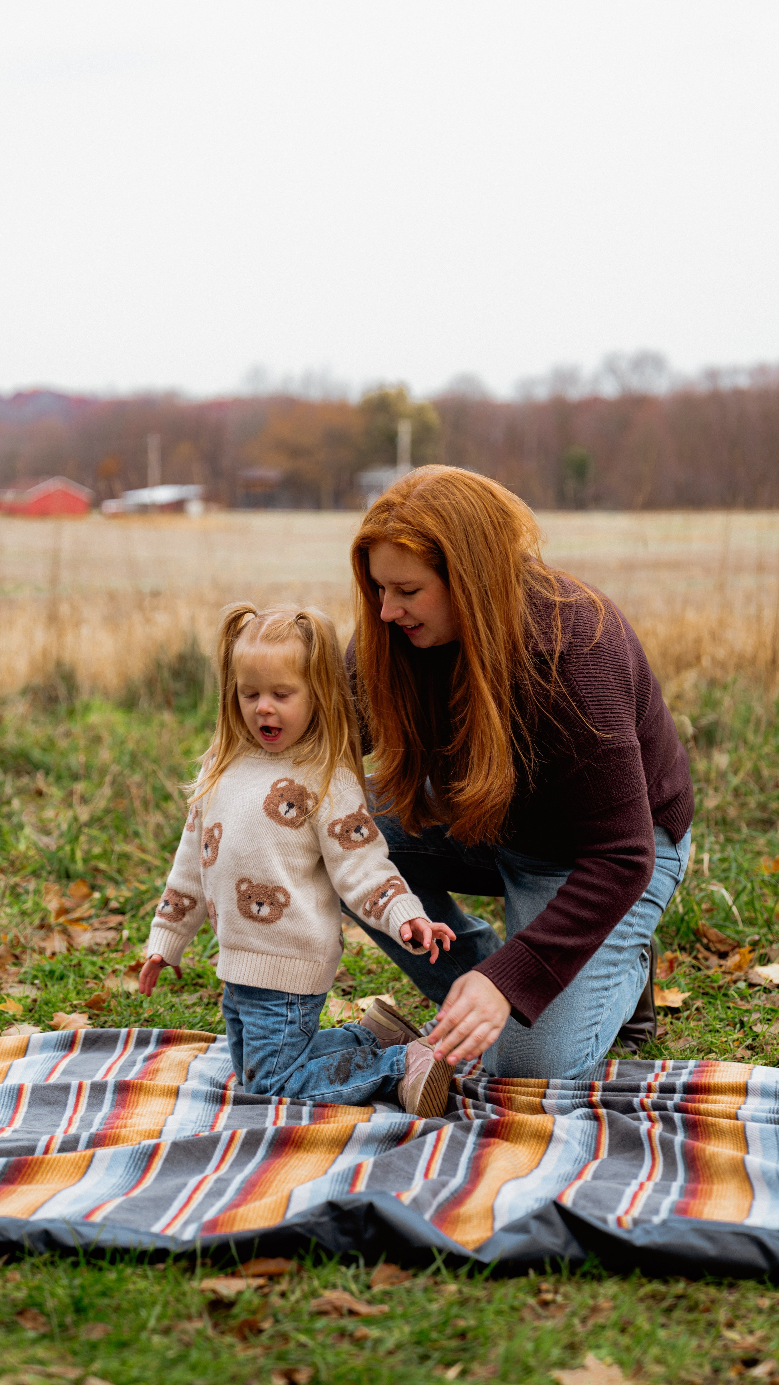 A mother and her young daughter outdoor on a fall day, kneeling on a striped blanket on the grass, with trees and field in the background.