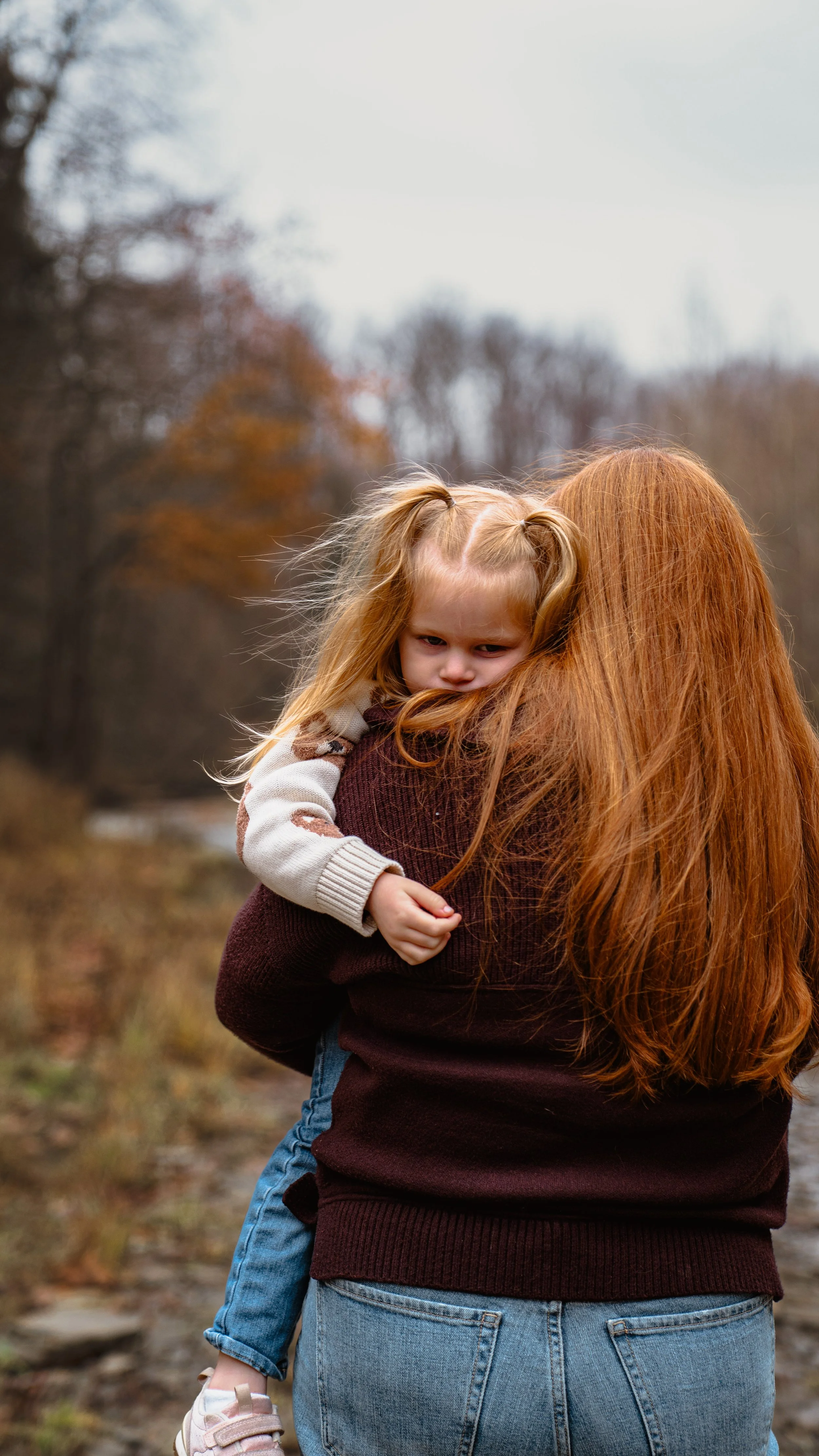 A woman with long red hair holding a young girl with blonde hair in a ponytail. The girl looks upset and is hugging the woman's shoulder outdoors in a fall setting with trees.