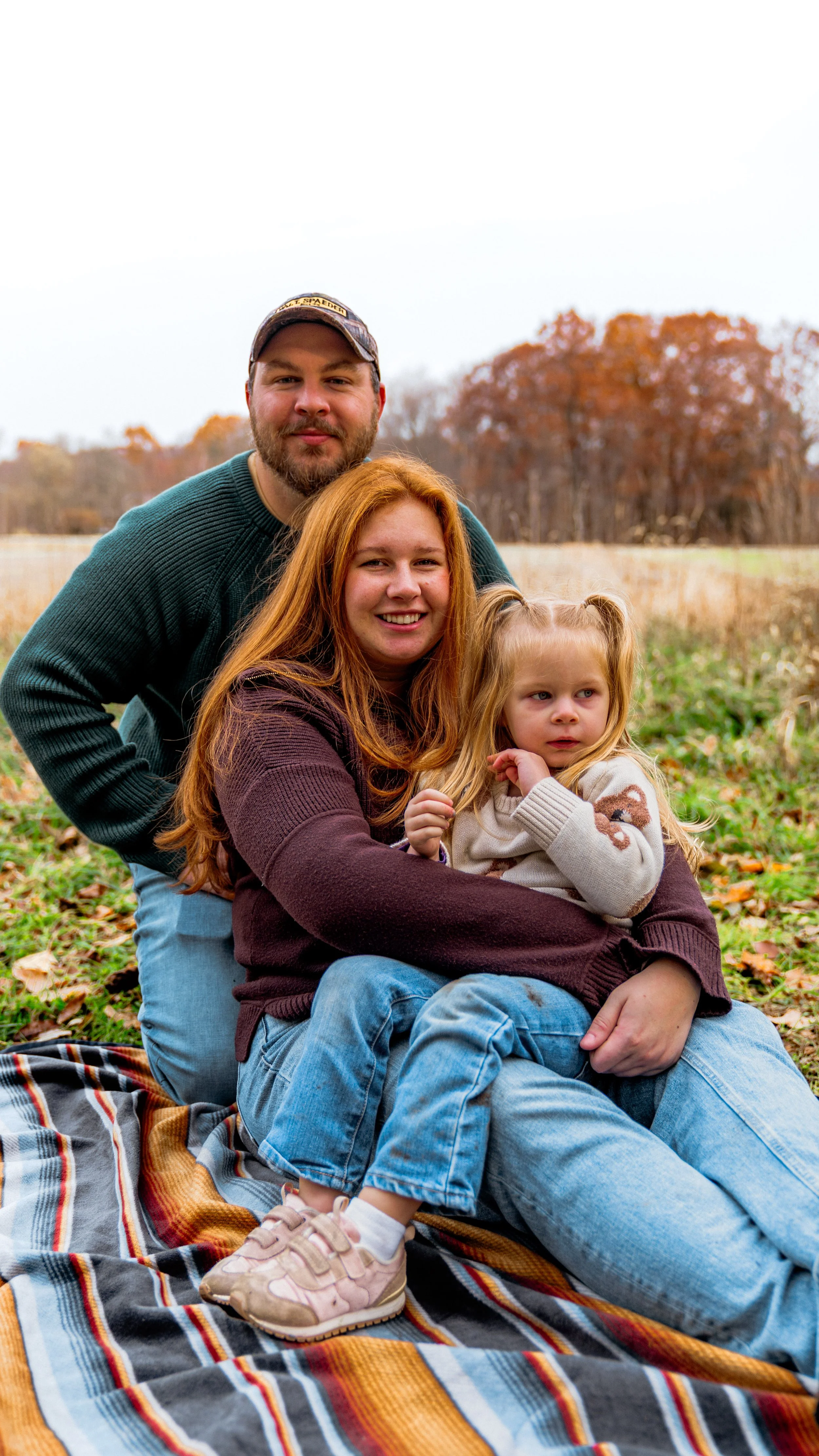 A family of three enjoying an outdoor fall day. The mother has long red hair and is sitting on a blanket, holding her young daughter with blonde pigtails. The father, with a beard, is standing behind them, leaning forward. The background features tre
