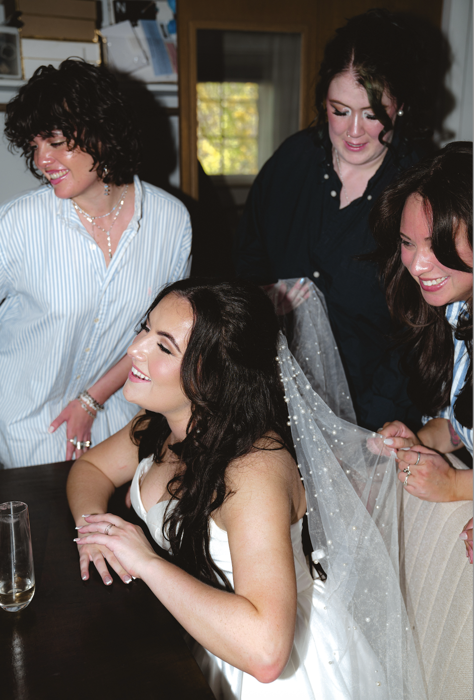 A bride with long dark hair, wearing a white wedding dress and veil, is smiling with her eyes closed, surrounded by three women helping her prepare for her wedding in a room with a window and bookshelf.