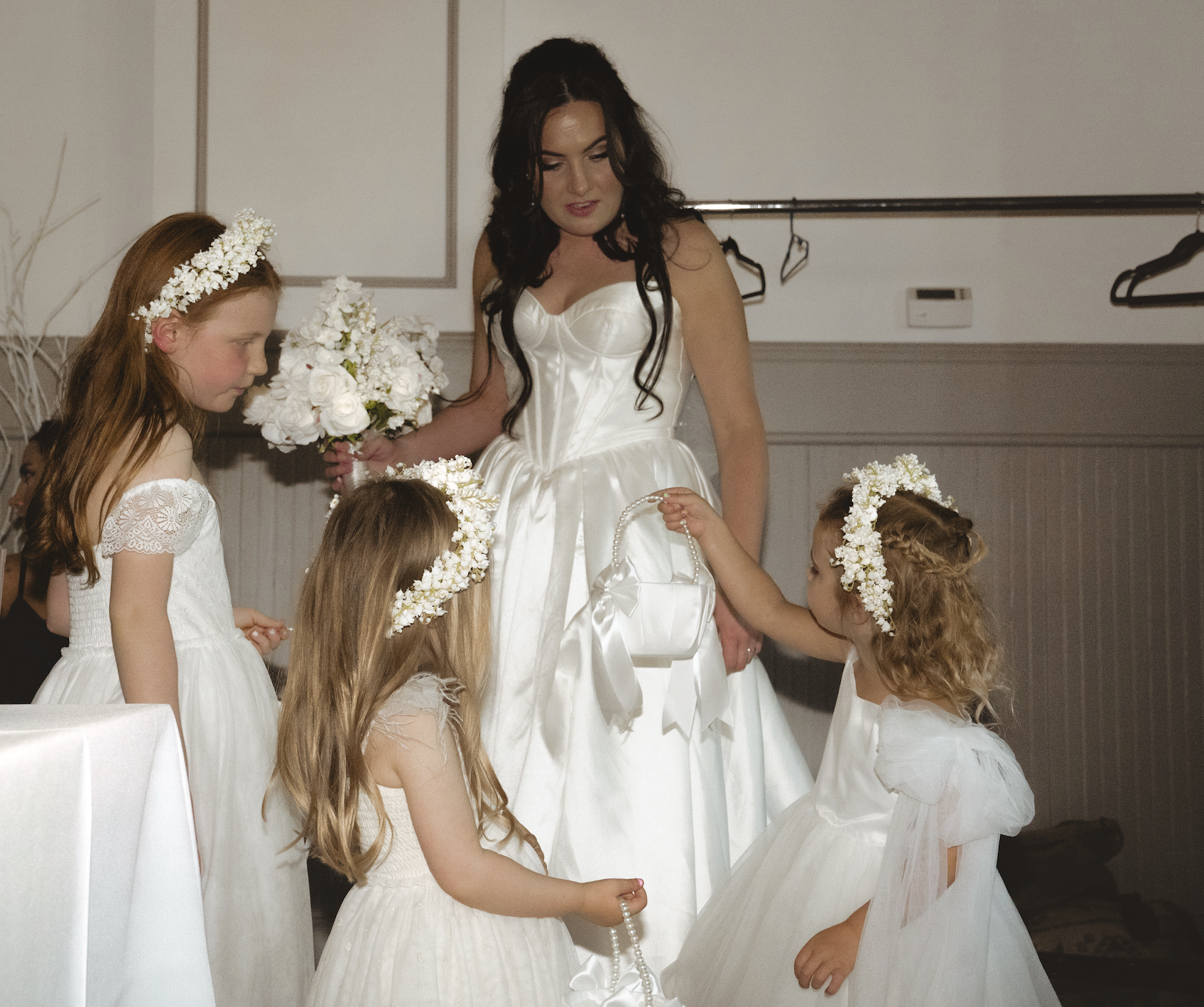 A bride in a white wedding dress holding a bouquet of flowers, surrounded by four young girls in white dresses and floral headbands, during a wedding event.
