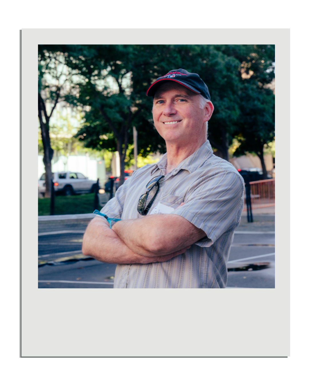 A man with gray hair wearing a baseball cap and sunglasses attached to his shirt, standing outdoors with arms crossed, smiling, in front of trees and parked cars.