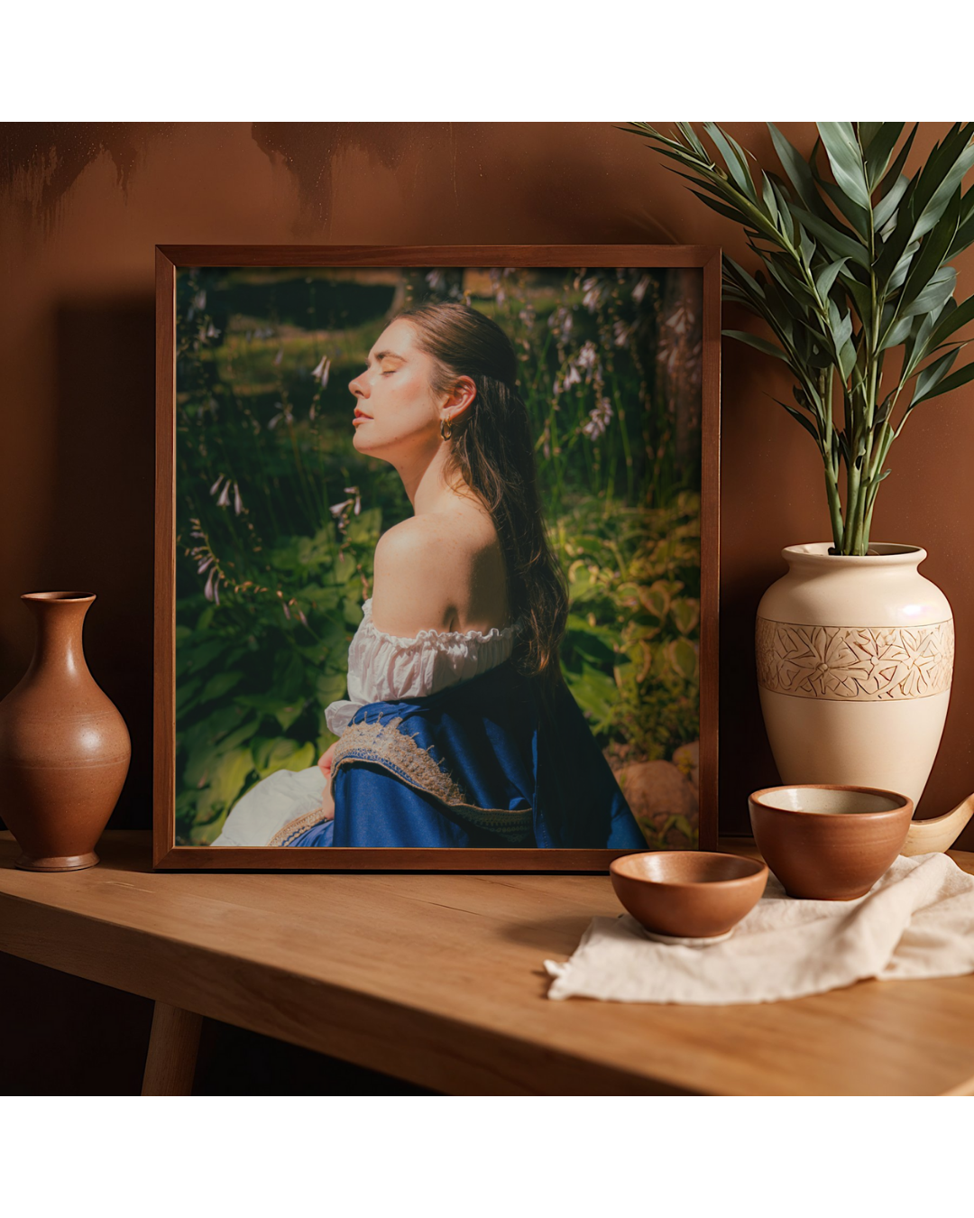 A framed photograph of a woman with long dark hair, wearing an off-shoulder top and a blue shawl, sitting outdoors with greenery in the background. The photo is placed on a wooden surface next to a tall plant in a decorative vase and several small pottery bowls.