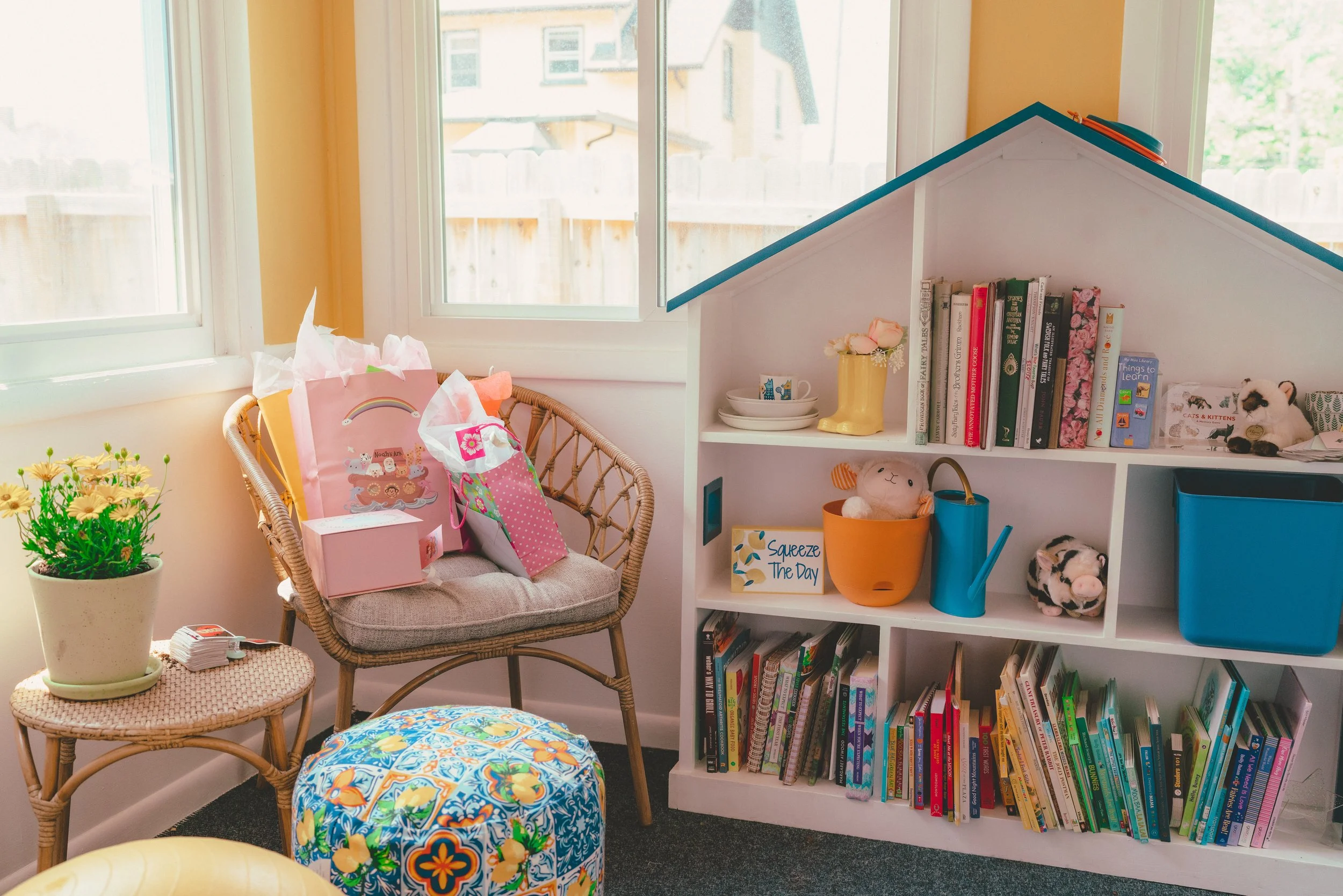 A cozy room corner with a rattan chair carrying gift bags, a small table with a potted plant, a colorful ottoman, and a bookshelf shaped like a house filled with books, toys, and decorative items.