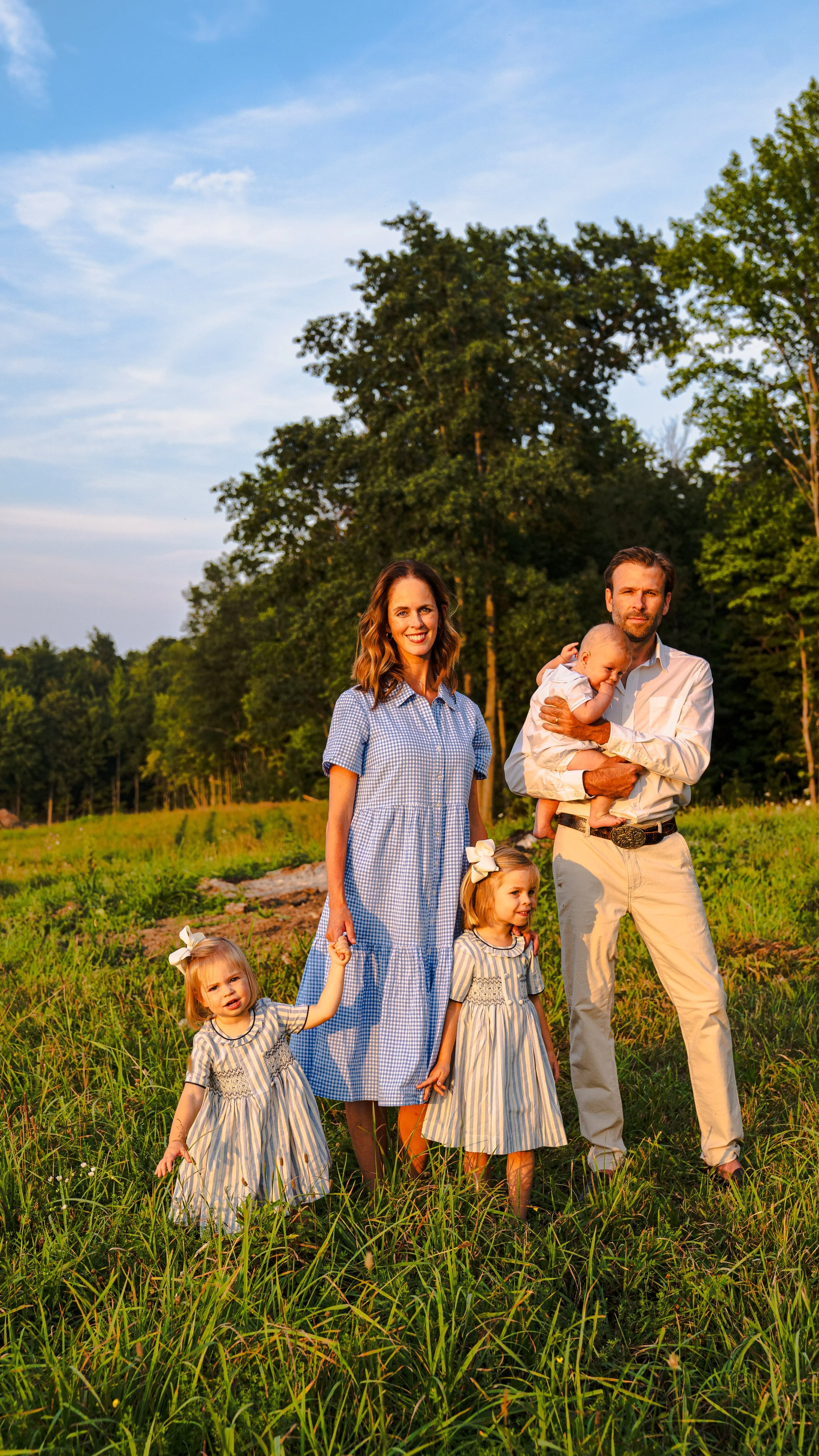 A family of five standing outdoors in a grassy field during sunset, with trees and blue sky in the background. The mother is wearing a blue checkered dress, and the father in a white shirt and beige pants. Two young girls in matching striped dresses are in front, and an infant is being held by the father.