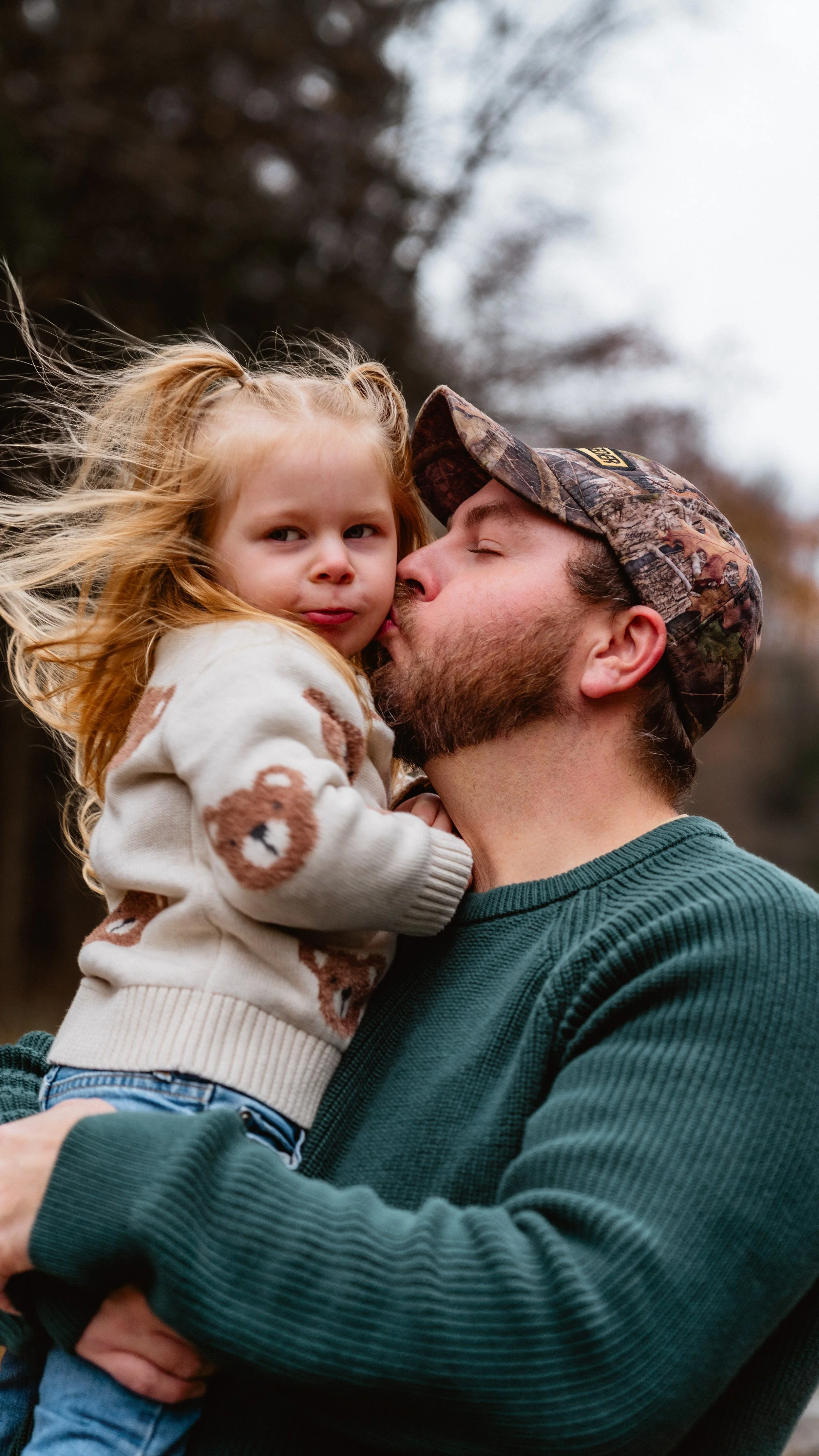 A man wearing a camouflage hat is kissing a young girl with strawberry blonde hair on the cheek. The girl is wearing a beige sweater with bear faces and has windblown hair. They are outdoors with a blurred background of trees and cloudy sky.