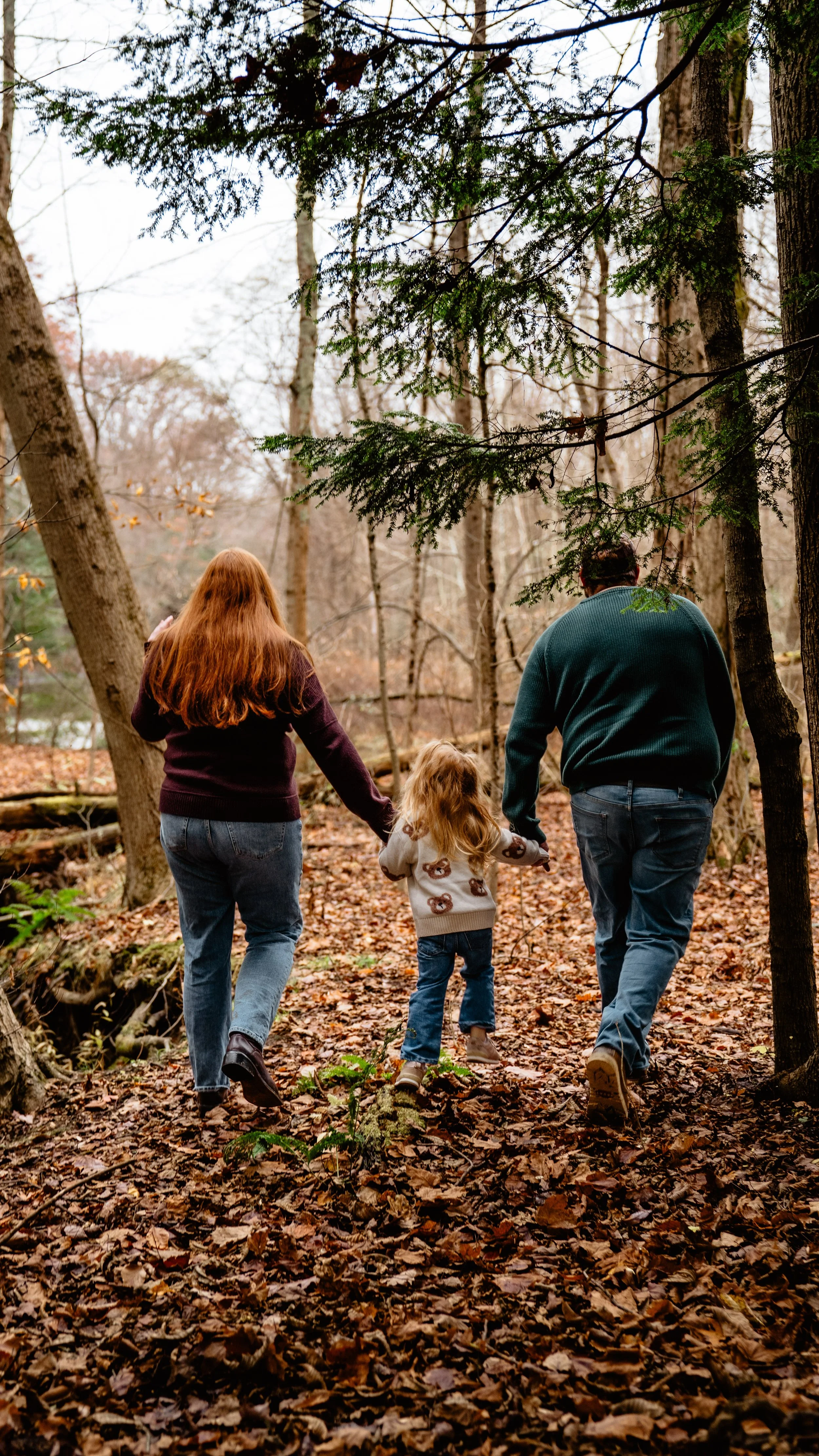 A family of three walking through a wooded forest during autumn, holding hands.