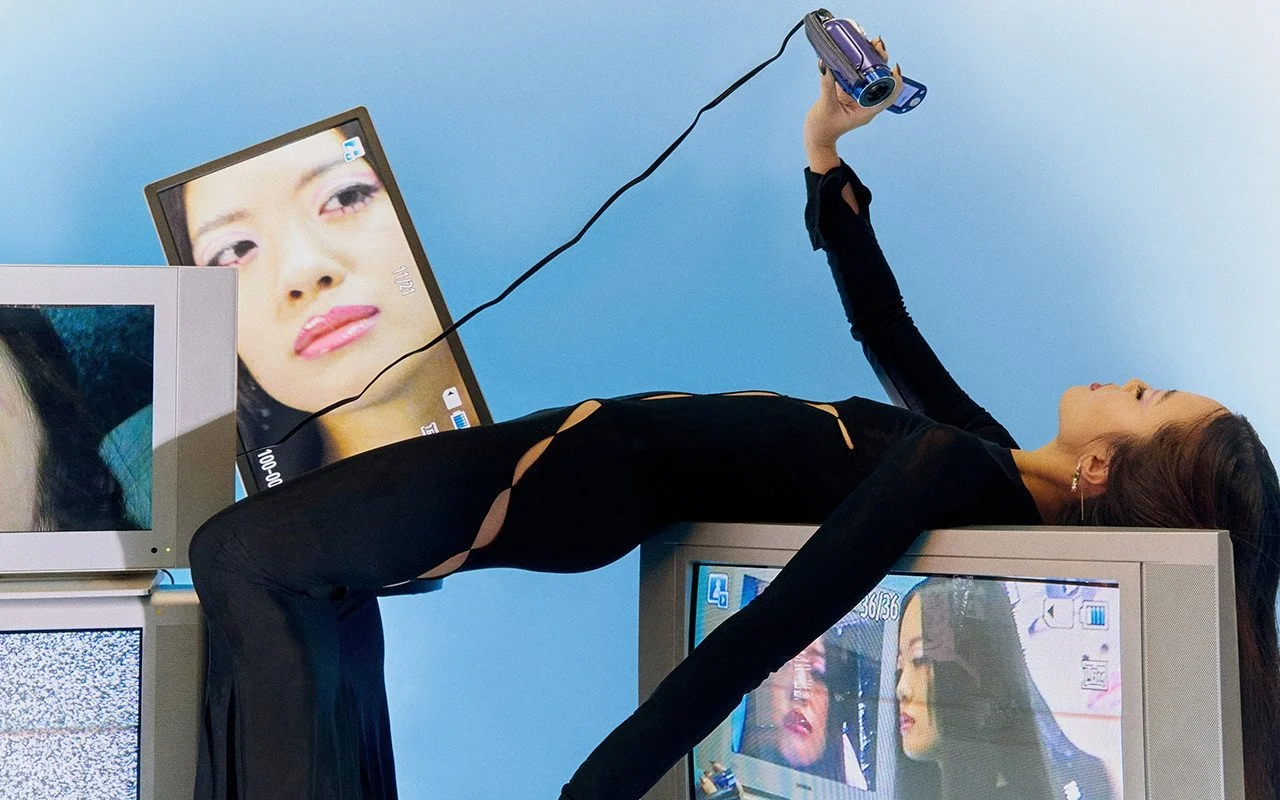 A woman lays on top of a CRT TV with an old-school  cam corder recording herself with the images appearing on the screen.