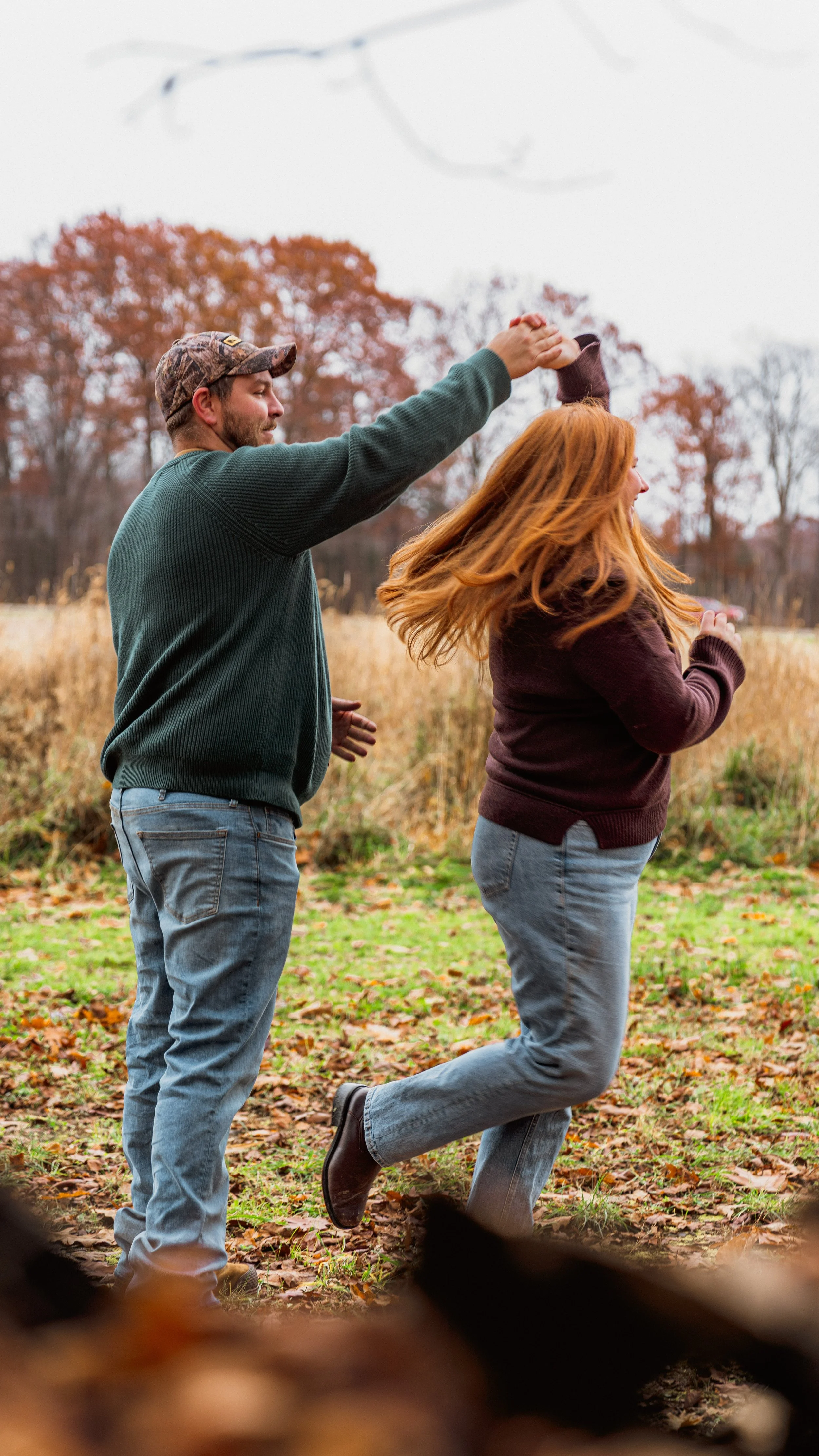 A man spinning a woman around in a park during fall, with trees and fallen leaves in the background.
