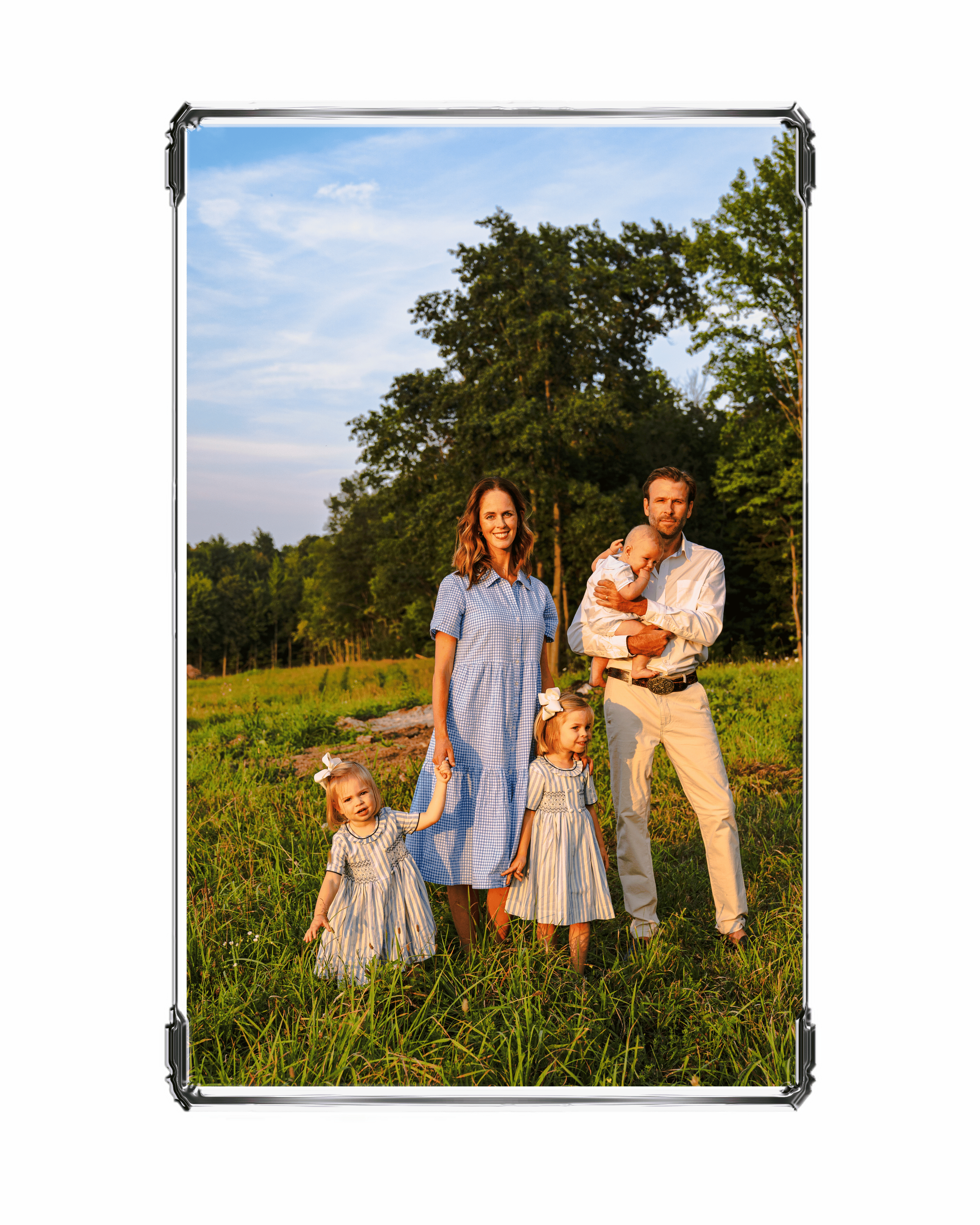 A family of five standing in a grassy field during sunset with tall trees and a blue sky in the background.