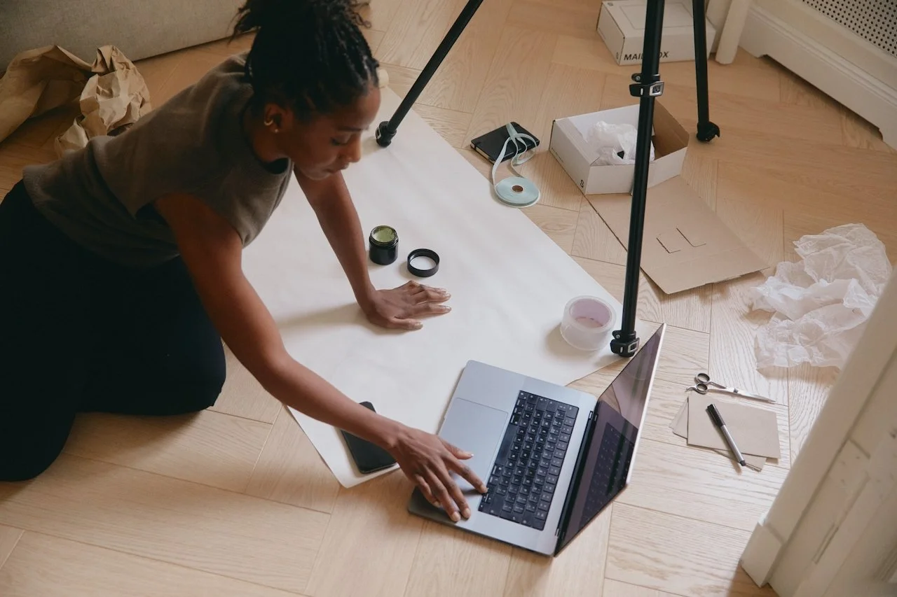 A woman sits on the floor with her computer and products underneath a tripod doing product photography.
