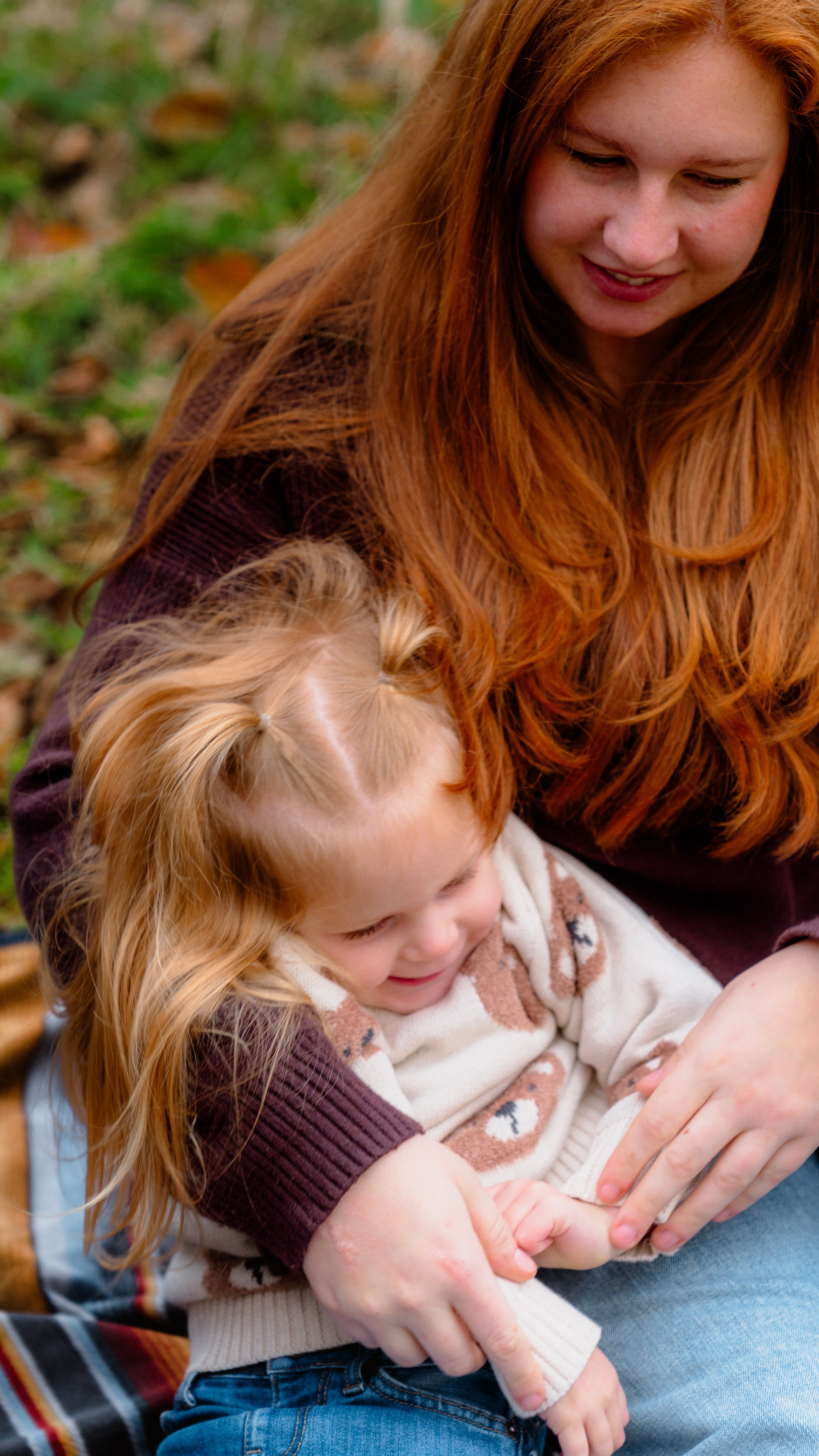 A woman with long red hair and a young girl with blonde hair are sitting outdoors on a fall day. The woman is gently holding the girl in her lap, and both are smiling, enjoying each other's company.